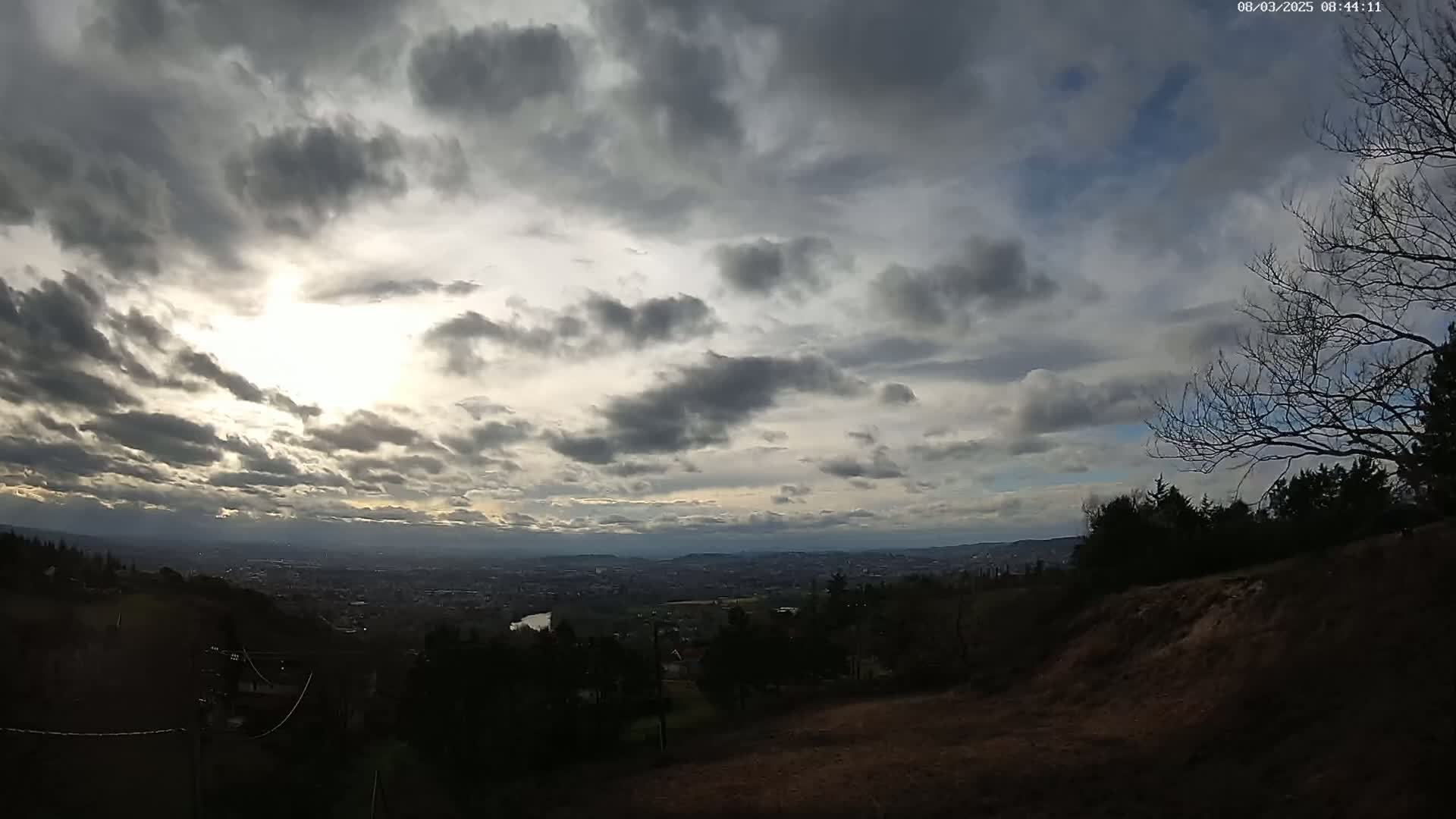 A view from a hilltop reveals a distant city under a mostly cloudy sky, with the sun faintly visible through the breaks in the clouds.
