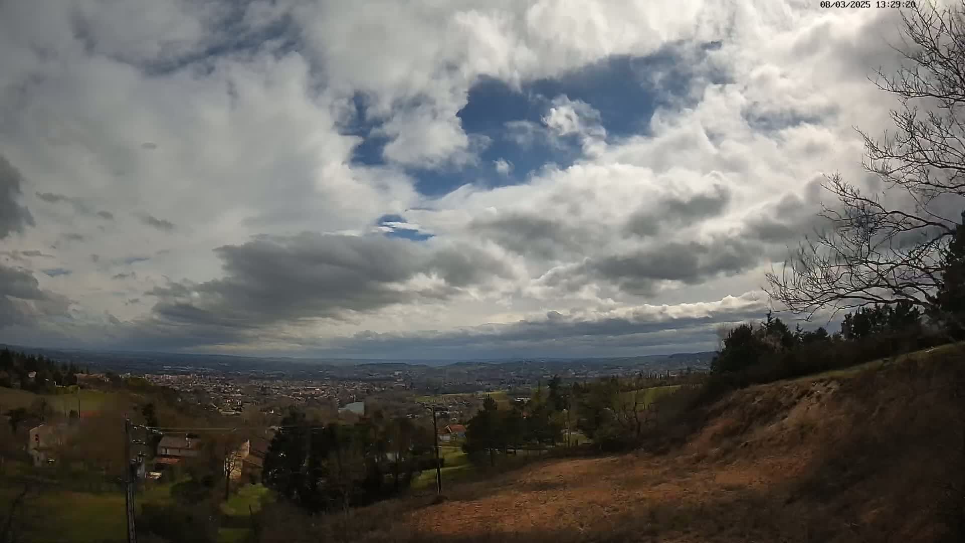 A hilly landscape with sparsely wooded areas overlooks a distant city under a partly cloudy sky with patches of blue.