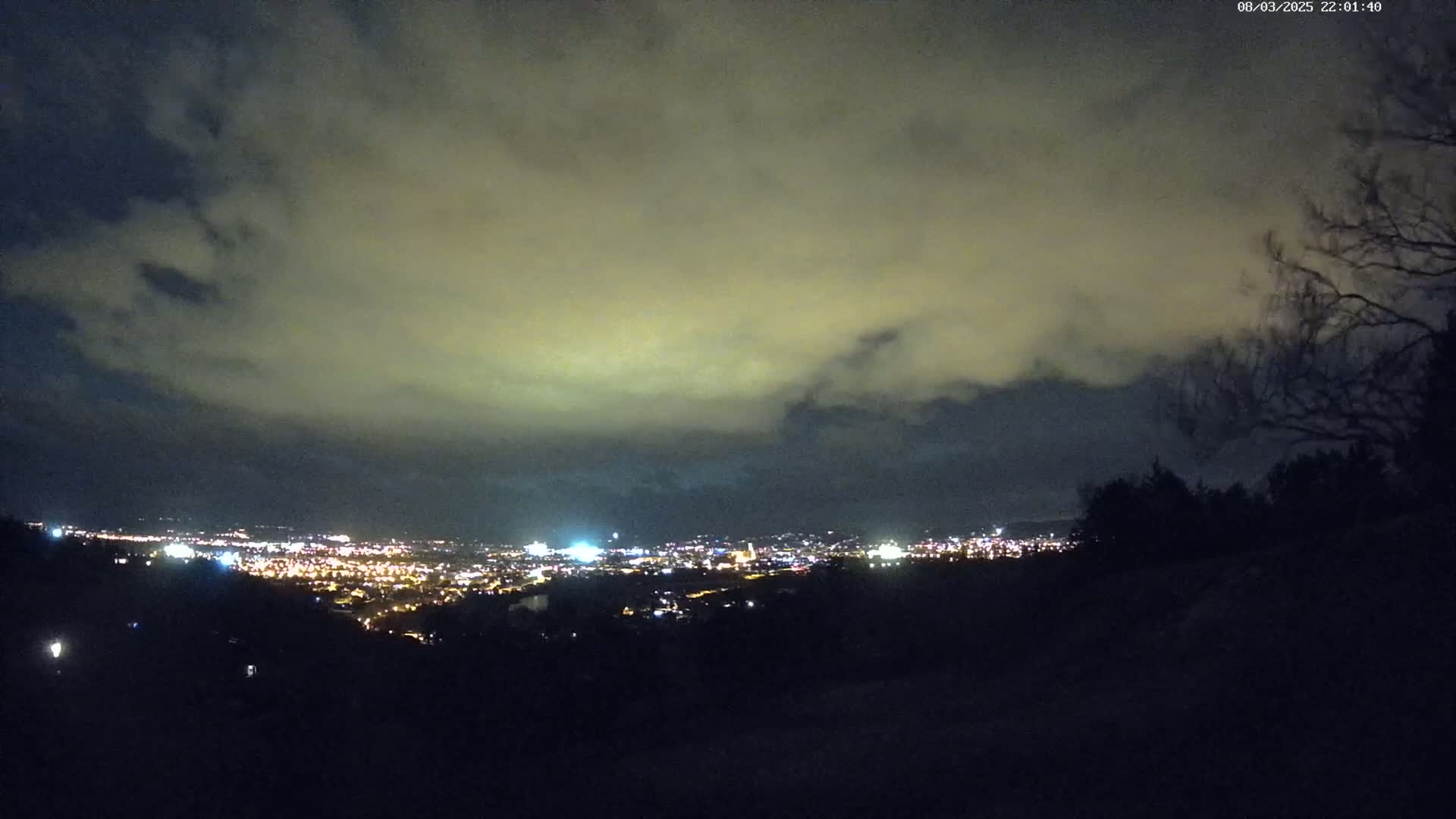 A nighttime view of a city's lights spread across a valley under a cloudy, yellowish sky.