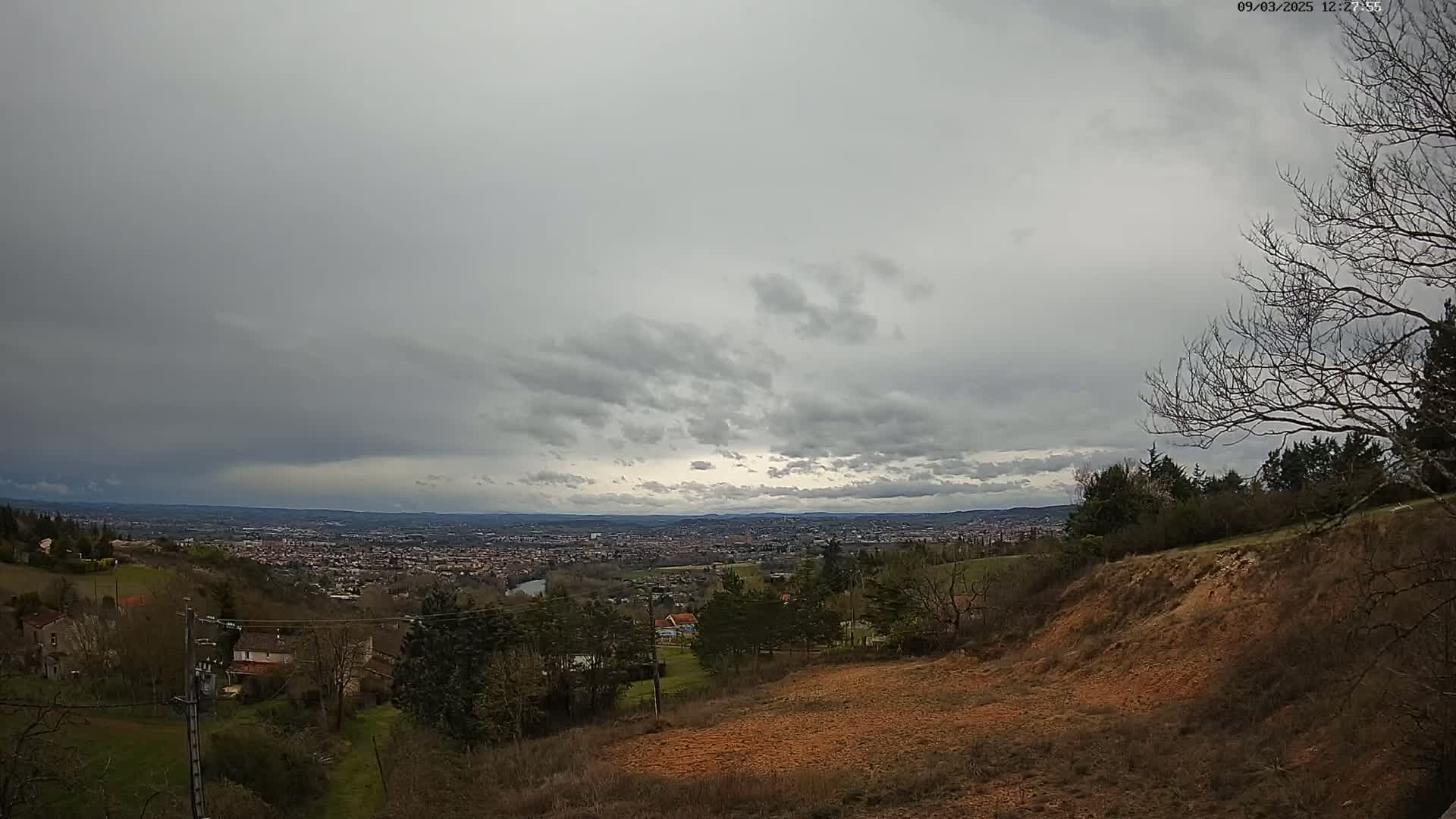 A sprawling view of a city under a heavily overcast sky is seen from a hilltop, with sparsely vegetated land in the foreground.