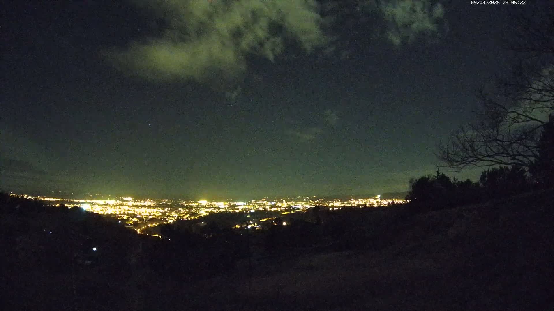 A nighttime view of a city's lights spread across a valley, partially obscured by a dark, tree-covered hillside under a cloudy sky.