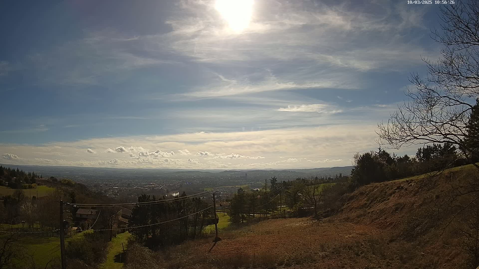 A sunny day with scattered clouds reveals a panoramic view of a sprawling city nestled in a valley, surrounded by rolling hills and sparsely wooded areas.