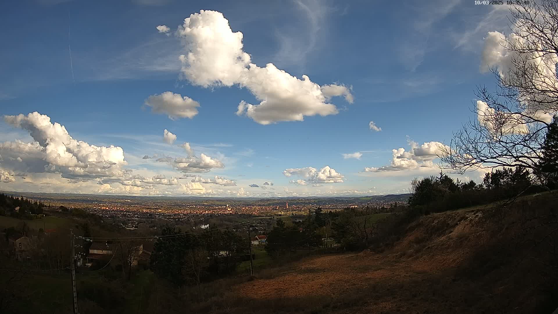 A hilly landscape under a partly cloudy blue sky overlooks a sprawling town in the distance.