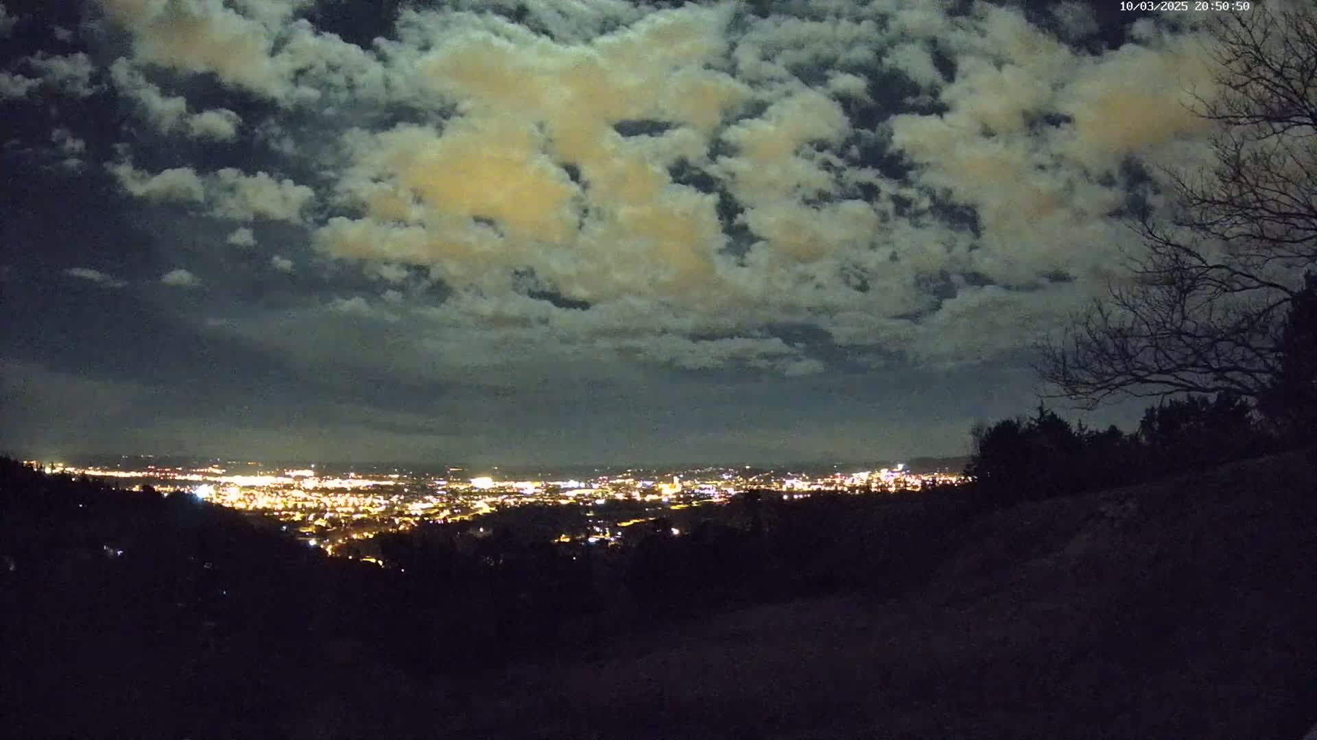 A nighttime view from a hilltop shows a city's lights spread across a valley under a sky filled with mostly cloudy conditions.