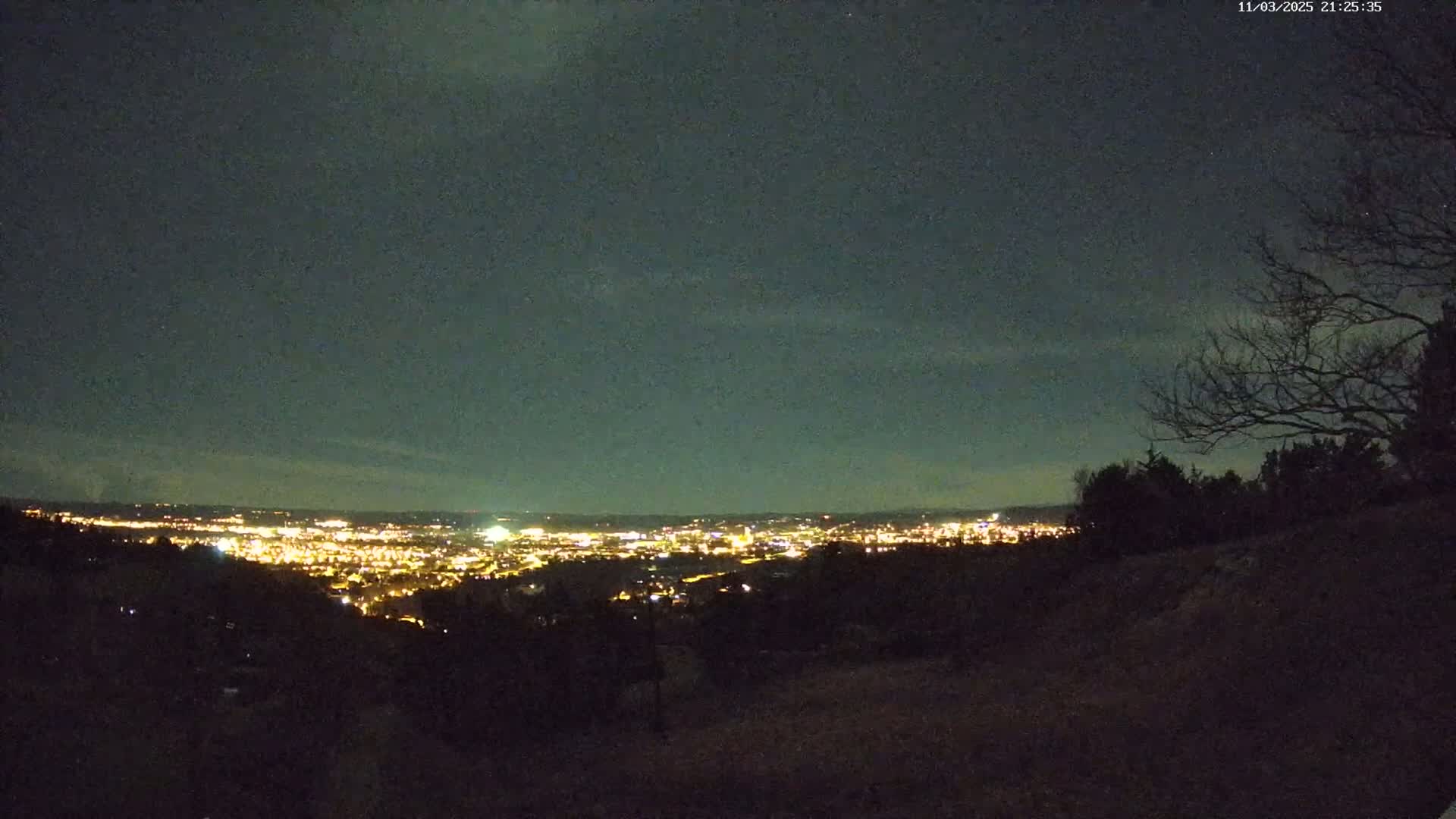 A nighttime view from a hilltop overlooking a city illuminated with lights under a mostly clear, dark sky.