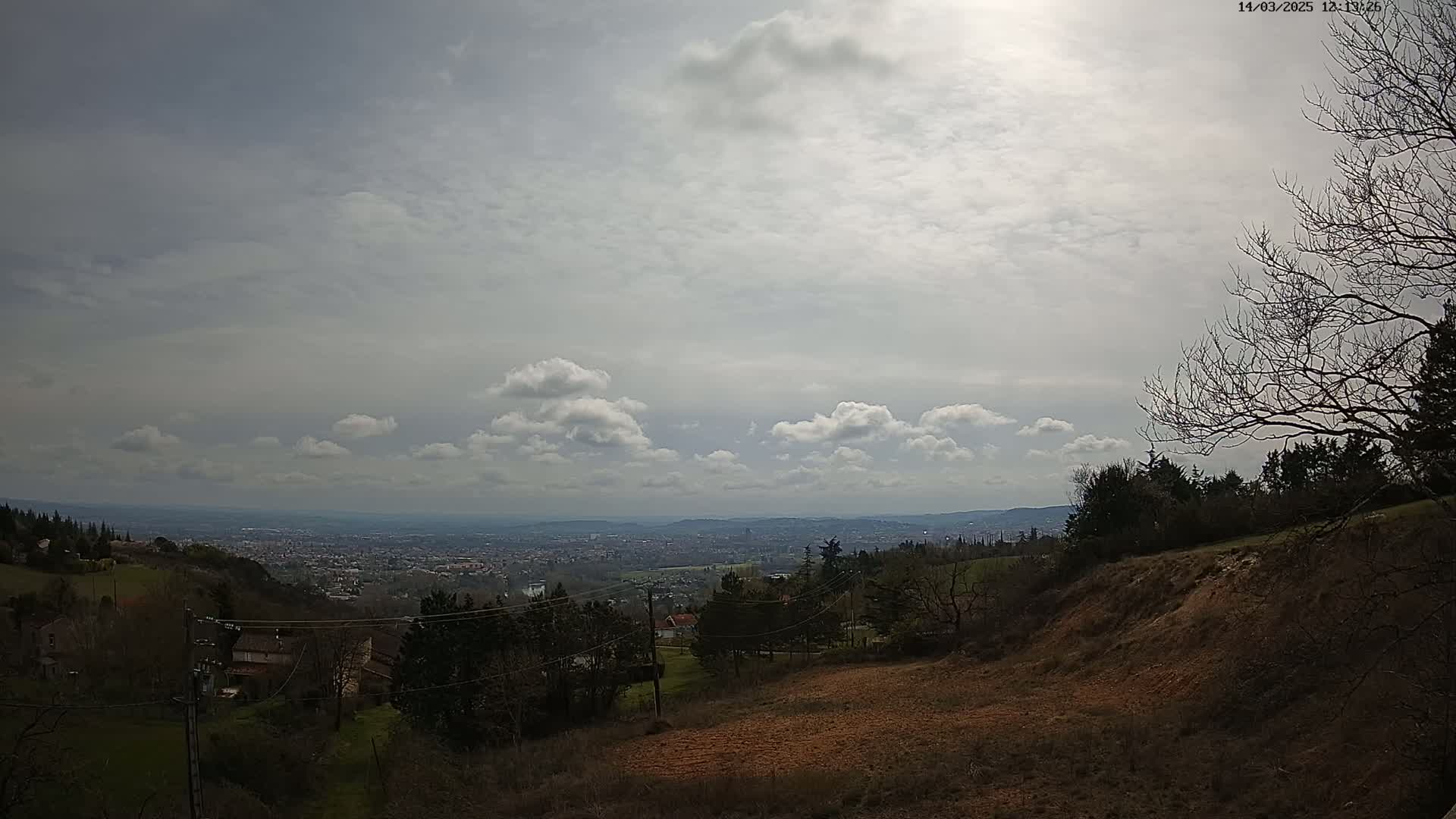 A partly cloudy sky overlooks a hilly landscape with sparsely wooded areas, houses, and a distant city visible in the background.