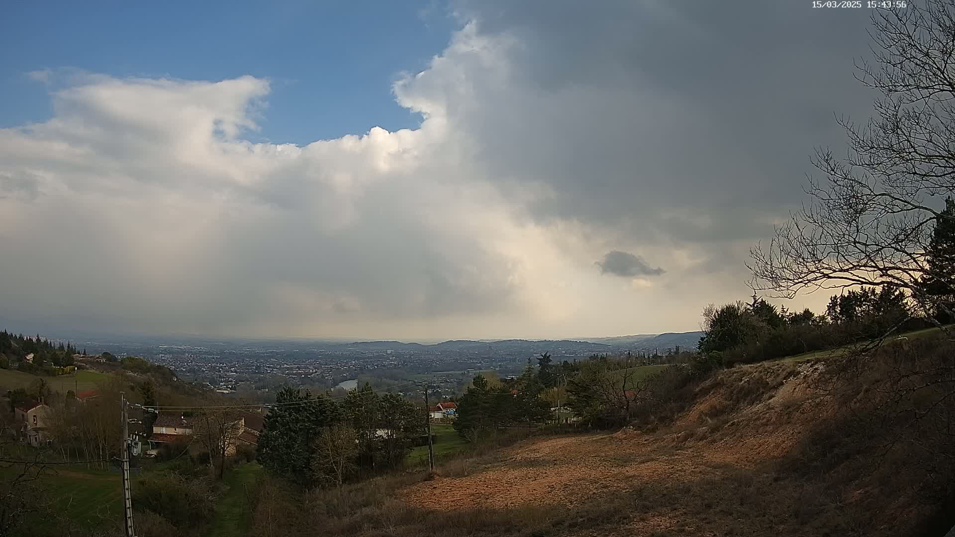 A hilly landscape with houses and trees under a partly cloudy sky, with a distant city visible in the background.