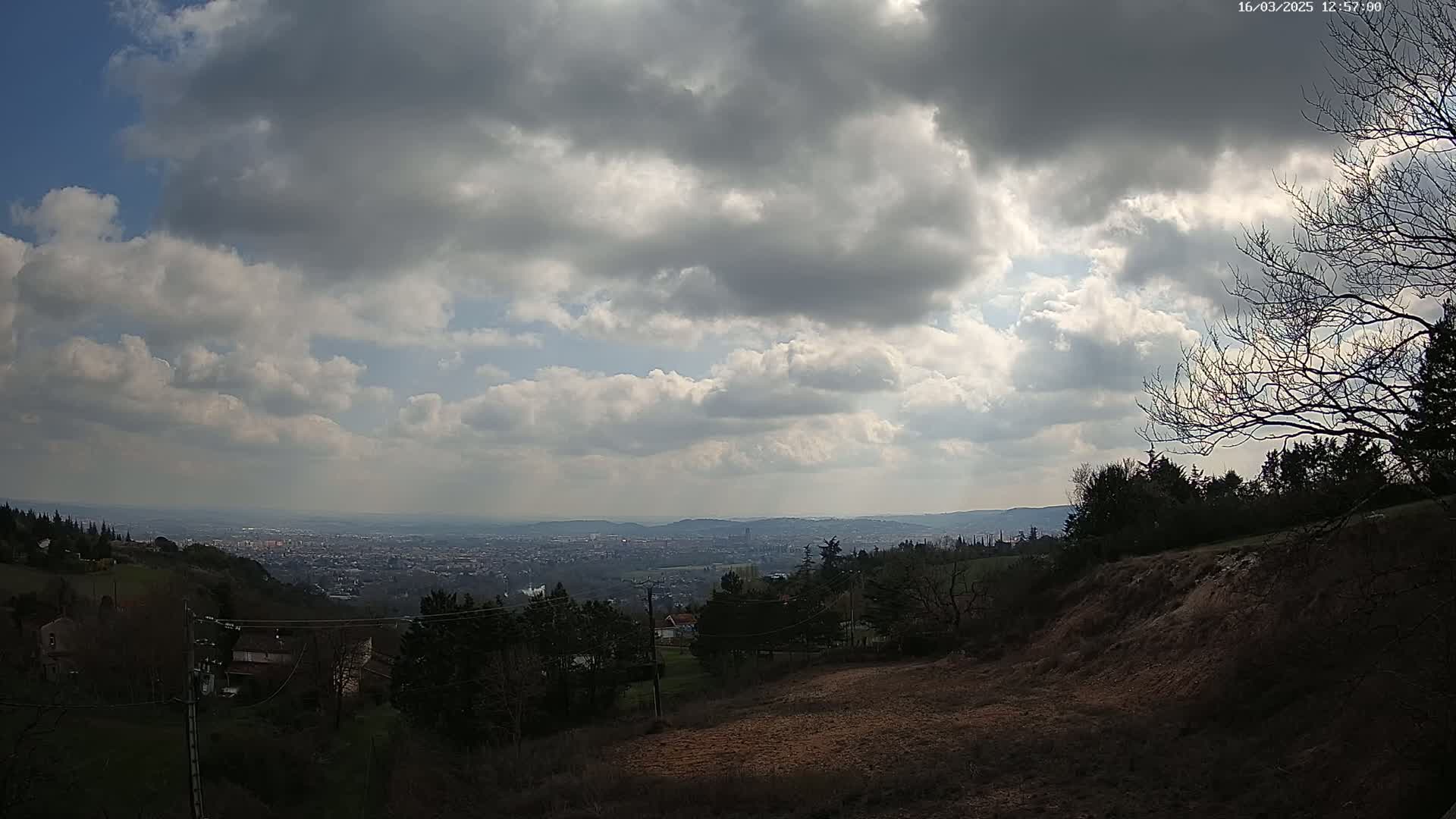 A partly cloudy sky overlooks a hilly landscape with distant city views and sparse, leafless trees in the foreground.