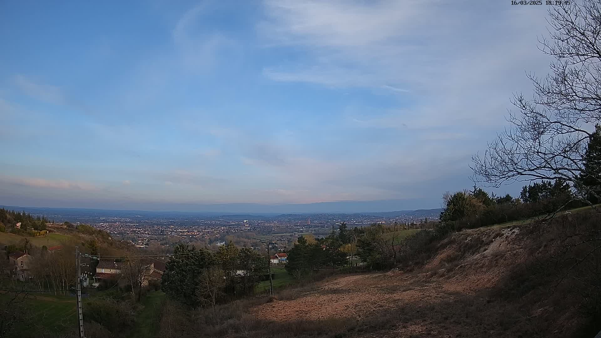 A sprawling city is visible in the distance under a mostly clear, light blue sky with a few wispy clouds, viewed from a hilly, sparsely wooded area.