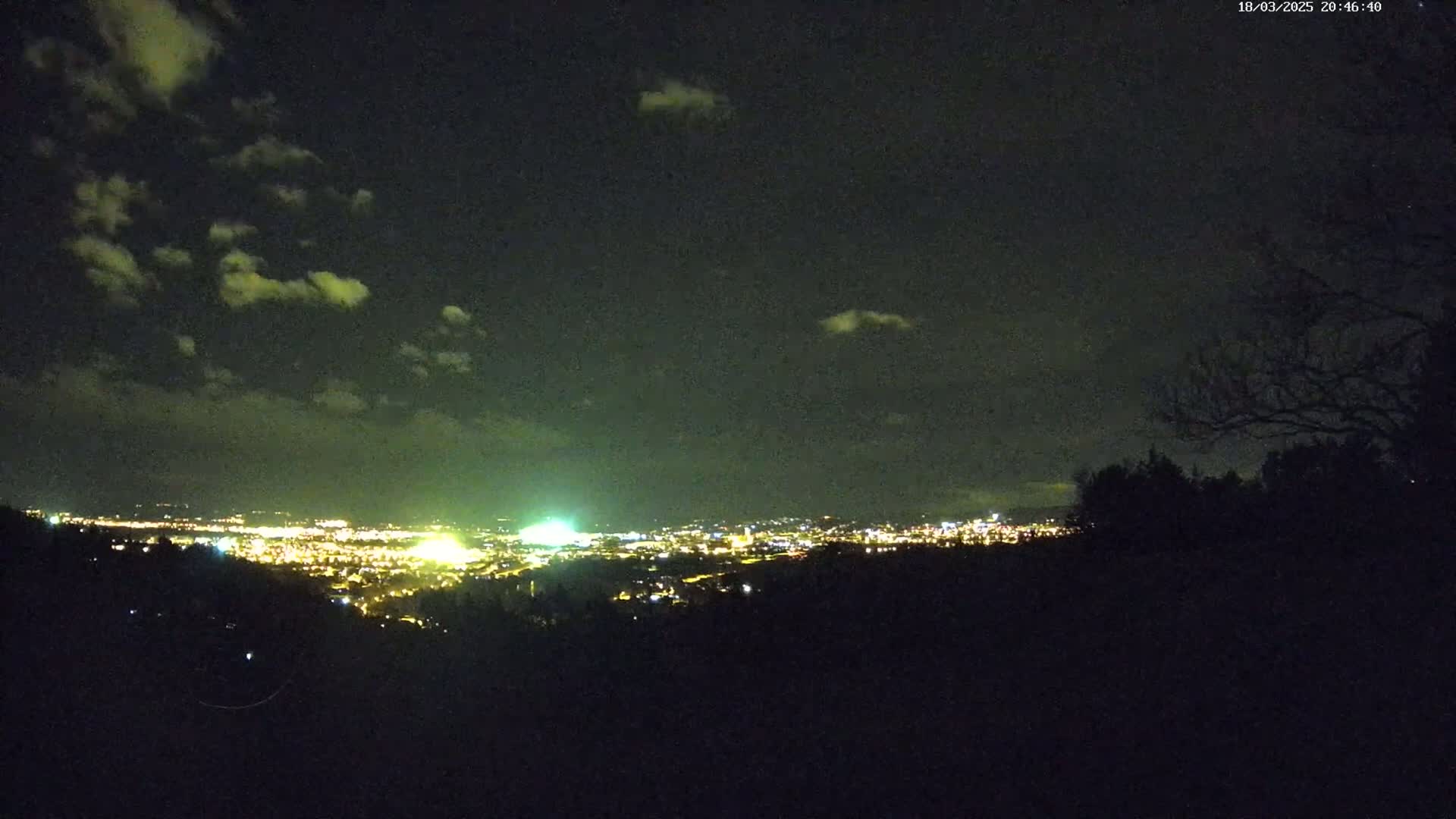 A nighttime view from a hilltop shows a city glowing with lights under a partly cloudy sky.