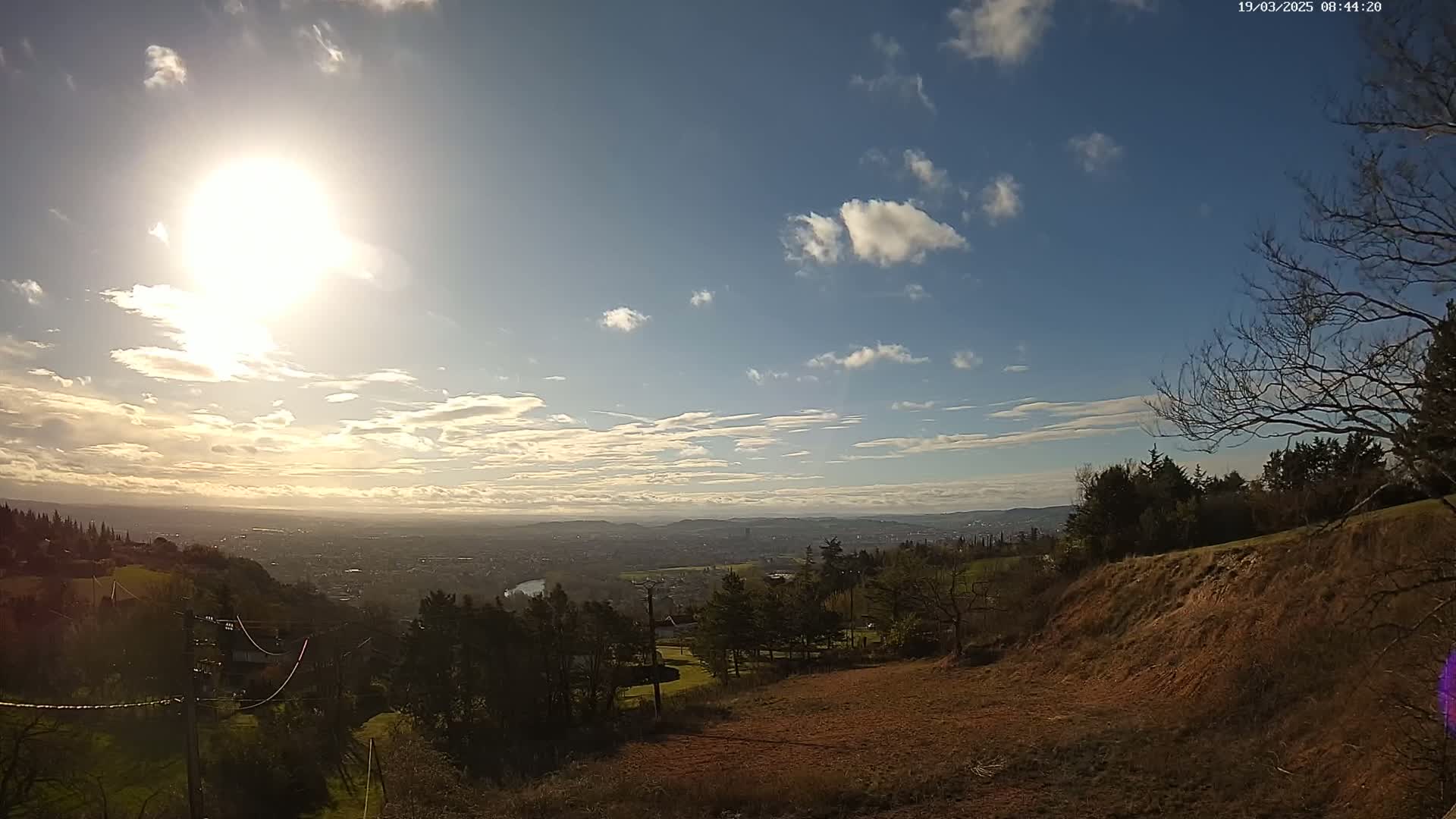 A sunny day with scattered clouds overlooks a vast city and landscape, viewed from a high vantage point with brown, barren land in the foreground.