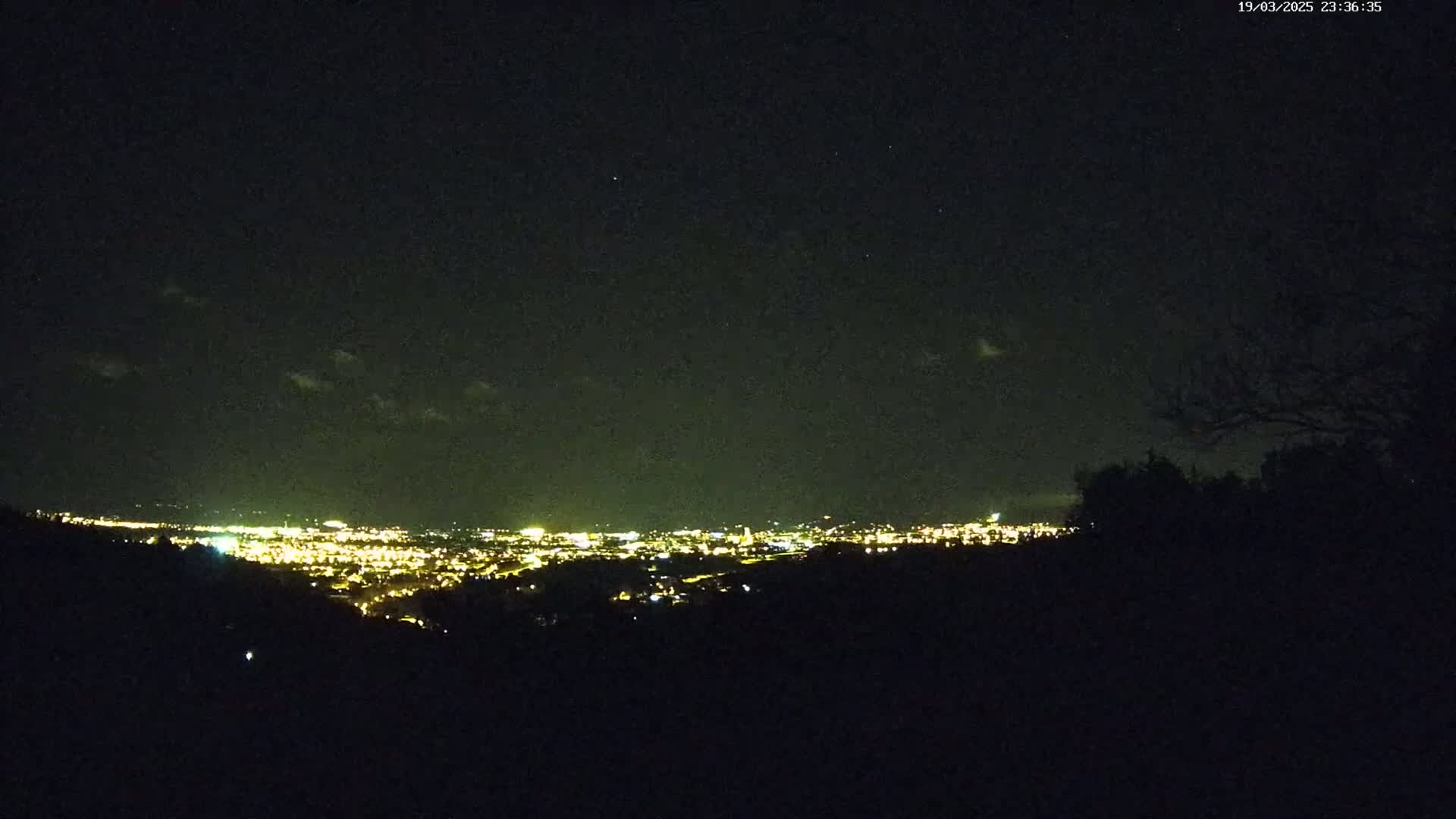 A nighttime view from a hilltop shows a distant city illuminated by many lights under a dark, mostly clear sky with a few clouds.