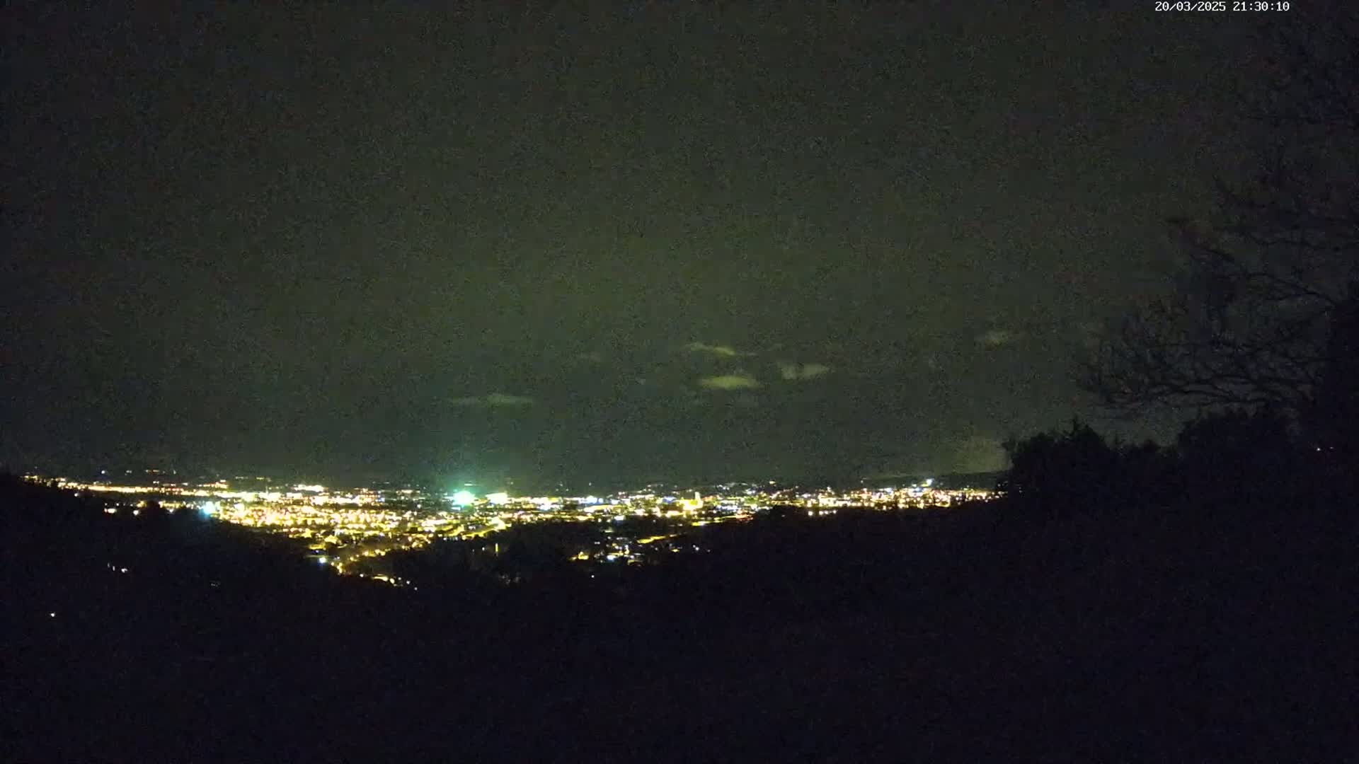 A nighttime view from a hilltop shows a sprawling city illuminated by countless lights under a dark, mostly clear sky with a few wispy clouds.