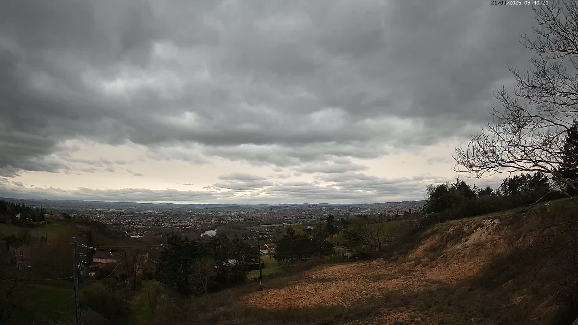 A sprawling view of a city nestled in a valley, under a cloudy sky, is seen from a hilltop overlooking sparsely wooded areas and a few houses.