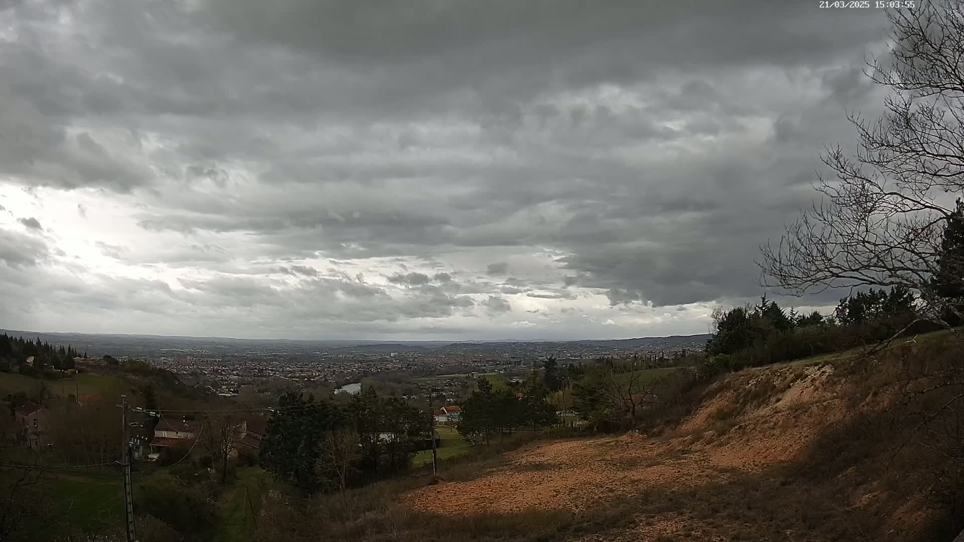 A sprawling cityscape is visible in the distance under a heavily overcast sky, viewed from a hillside with sparse vegetation and exposed earth.