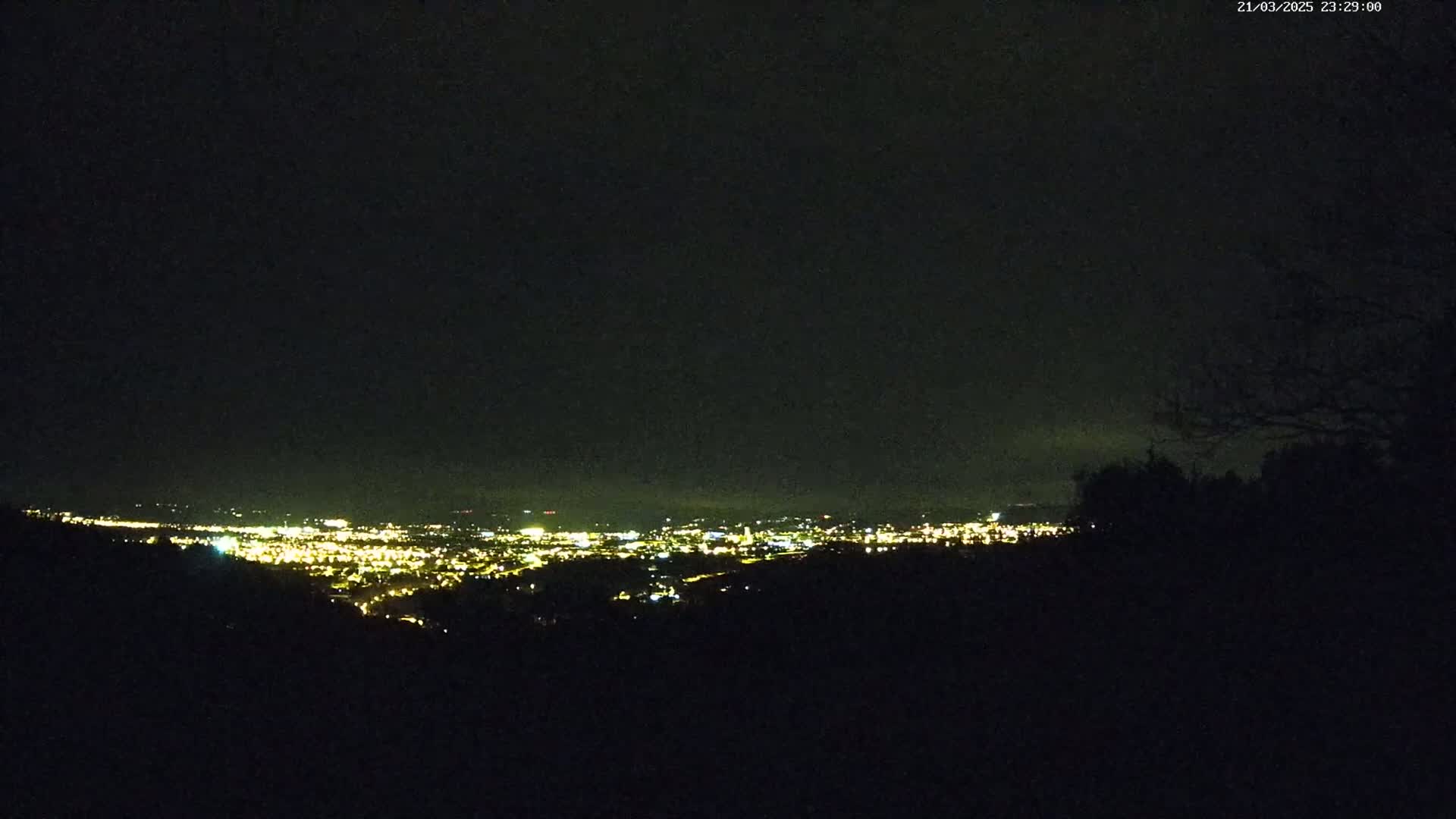 A nighttime view from a hilltop shows a distant city illuminated by many small lights under a dark, overcast sky.