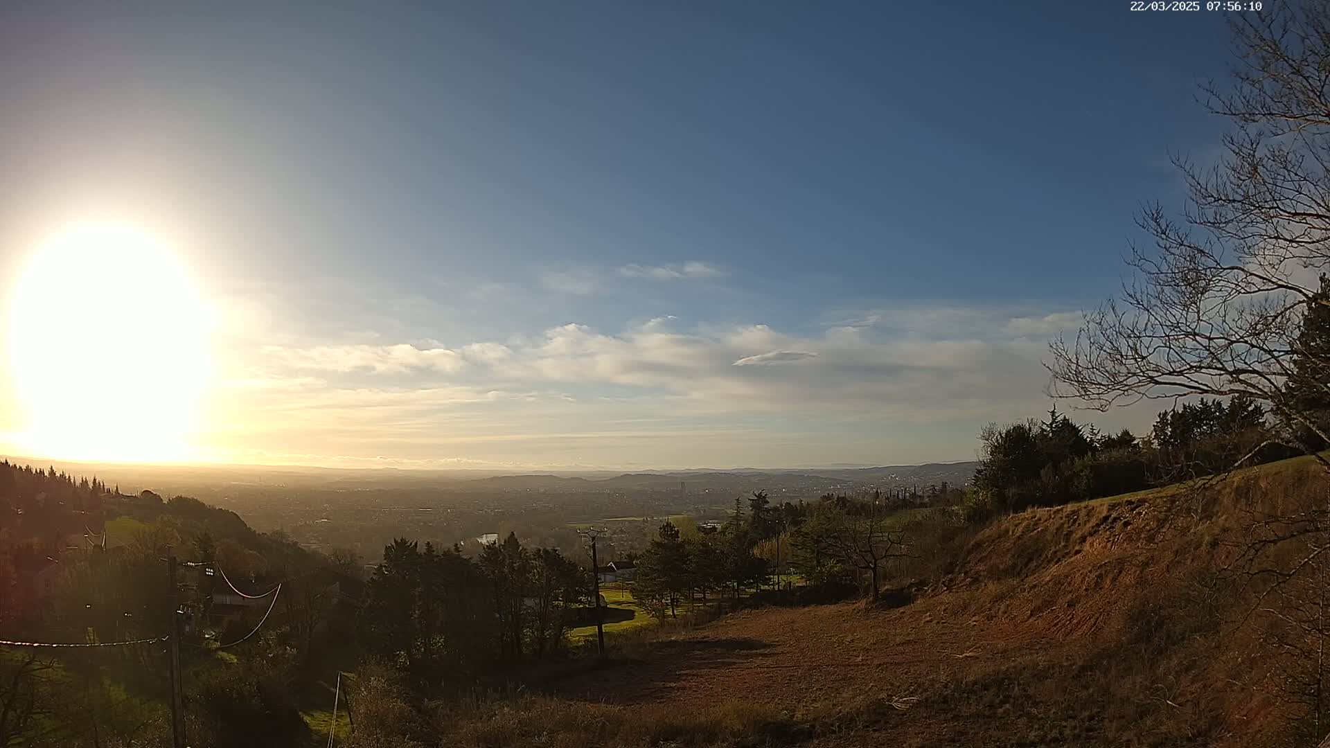 A sunny day with a few clouds reveals a distant city view from a hilltop overlooking a sparsely wooded area with a cleared section of land.