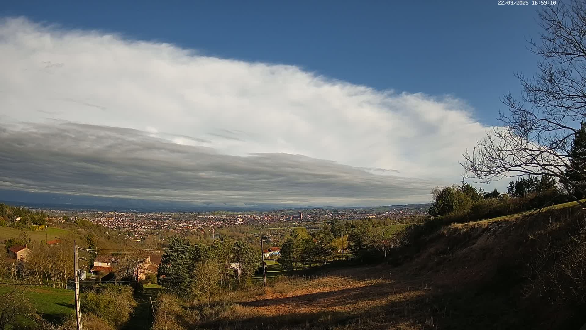 A mostly cloudy sky, with a layer of low, dark clouds over a distant city, viewed from a hillside with sparse trees and houses.