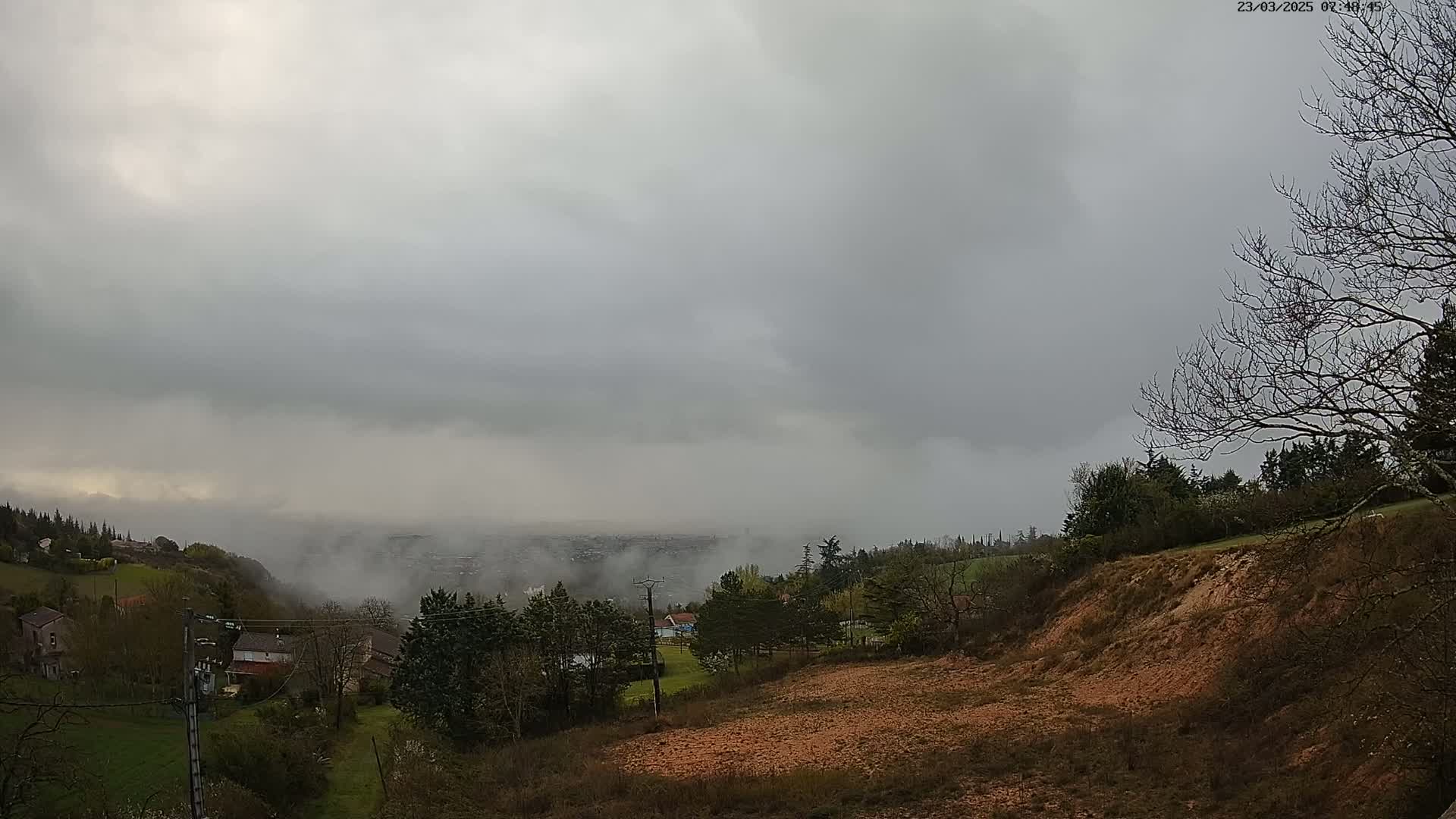 A low-lying fog obscures a distant city under a heavily overcast sky, while a hillside with leafless trees and sparse vegetation is visible in the foreground.