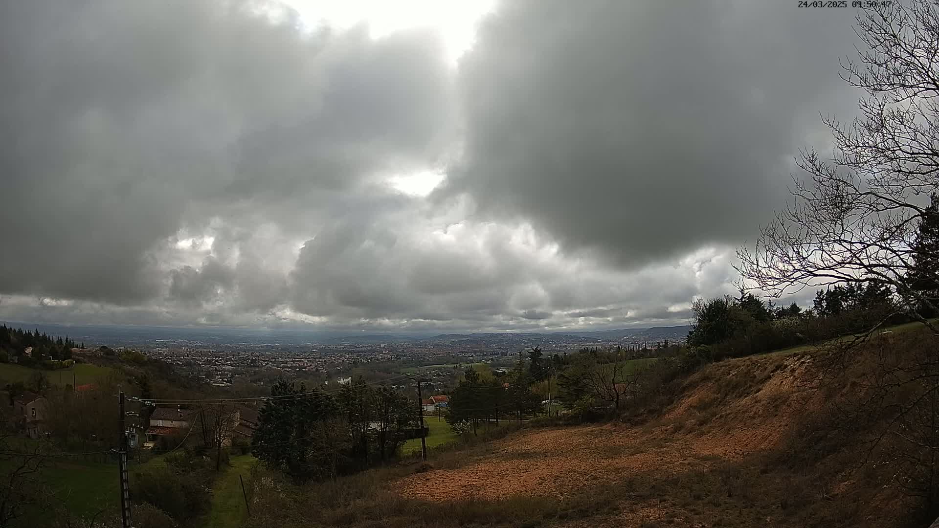 A view from a hillside shows a distant city under a heavily clouded sky.