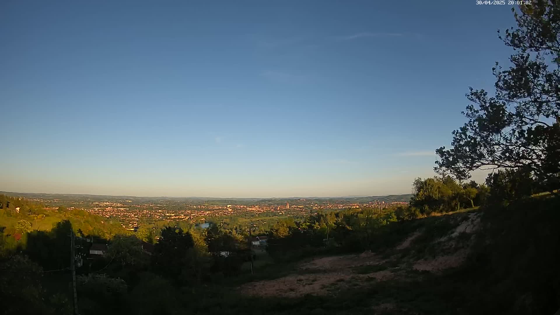 A sprawling city is visible in the distance under a clear blue sky, viewed from a hillside covered in green trees and shrubs.