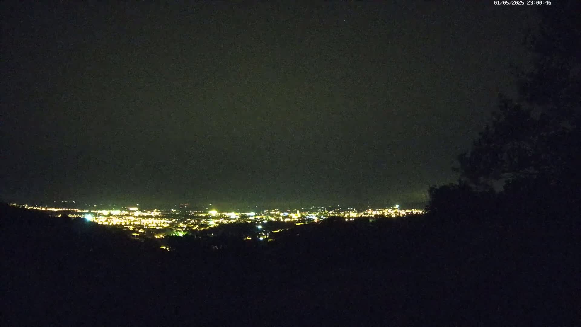 A nighttime view from a hilltop shows a distant city illuminated by lights under a dark, clear sky.