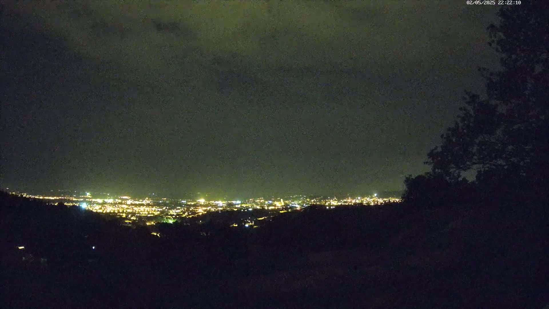 A nighttime view from a hilltop shows a distant city illuminated by numerous lights under a dark, cloudy sky.