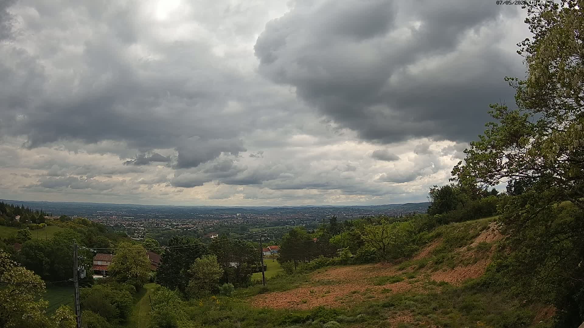 A sprawling city is visible in the distance under a heavily clouded sky, viewed from a hilltop covered in lush green vegetation and bare, reddish-brown earth.