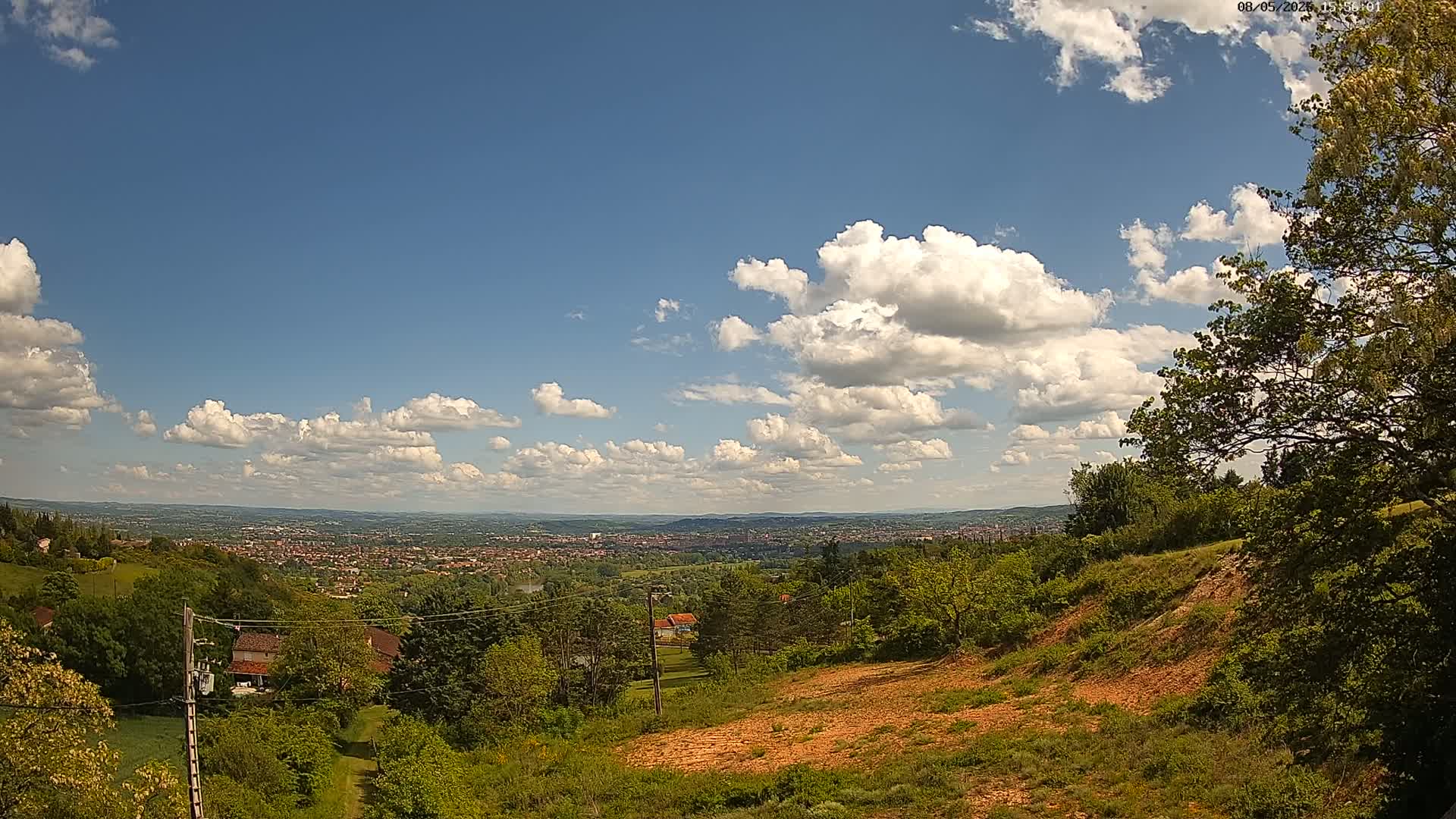 A partly cloudy day reveals a sprawling vista of a city nestled in a valley, viewed from a hilltop where lush green vegetation and exposed reddish-brown earth are present.