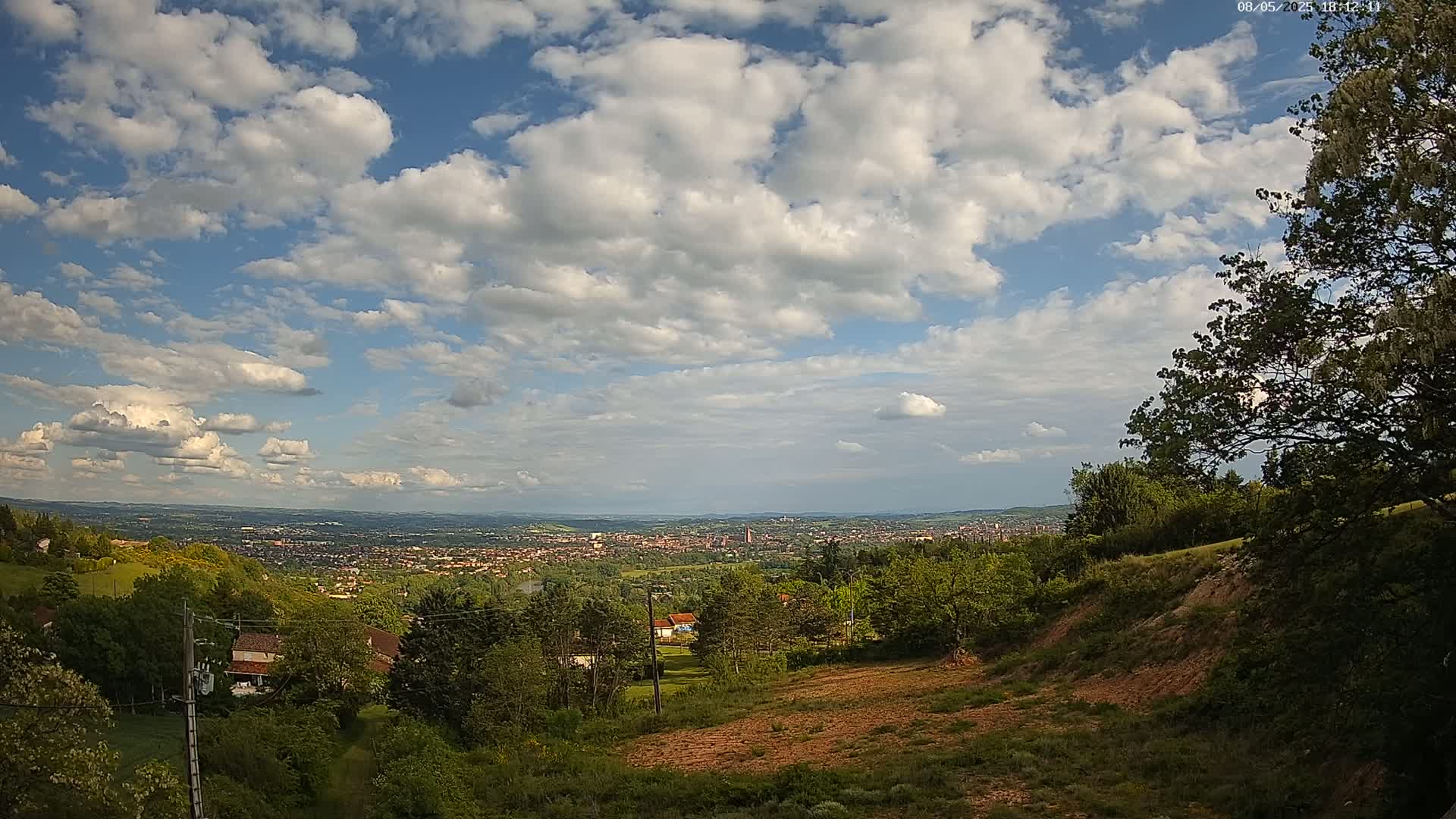 A hilly, green landscape under a partly cloudy sky overlooks a distant city.