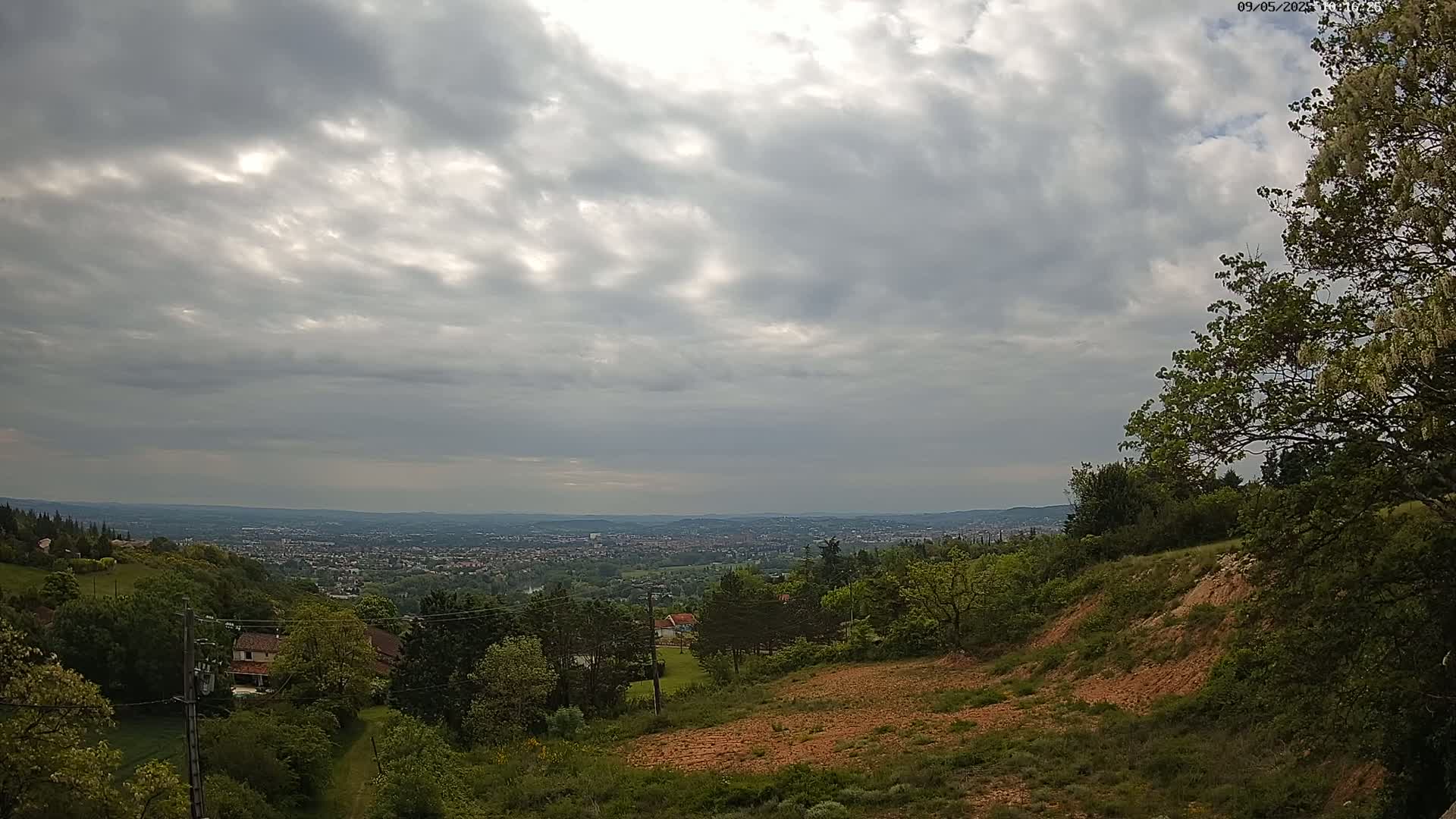 A hilly, wooded landscape overlooks a distant city under a mostly cloudy sky.