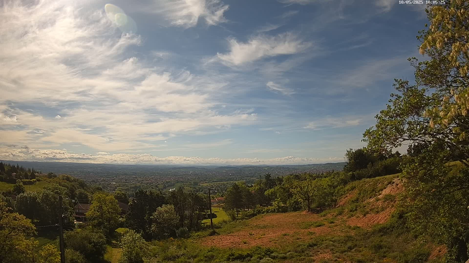 A hilly landscape with lush green trees and a distant view of a city under a partly cloudy, sunny sky.
