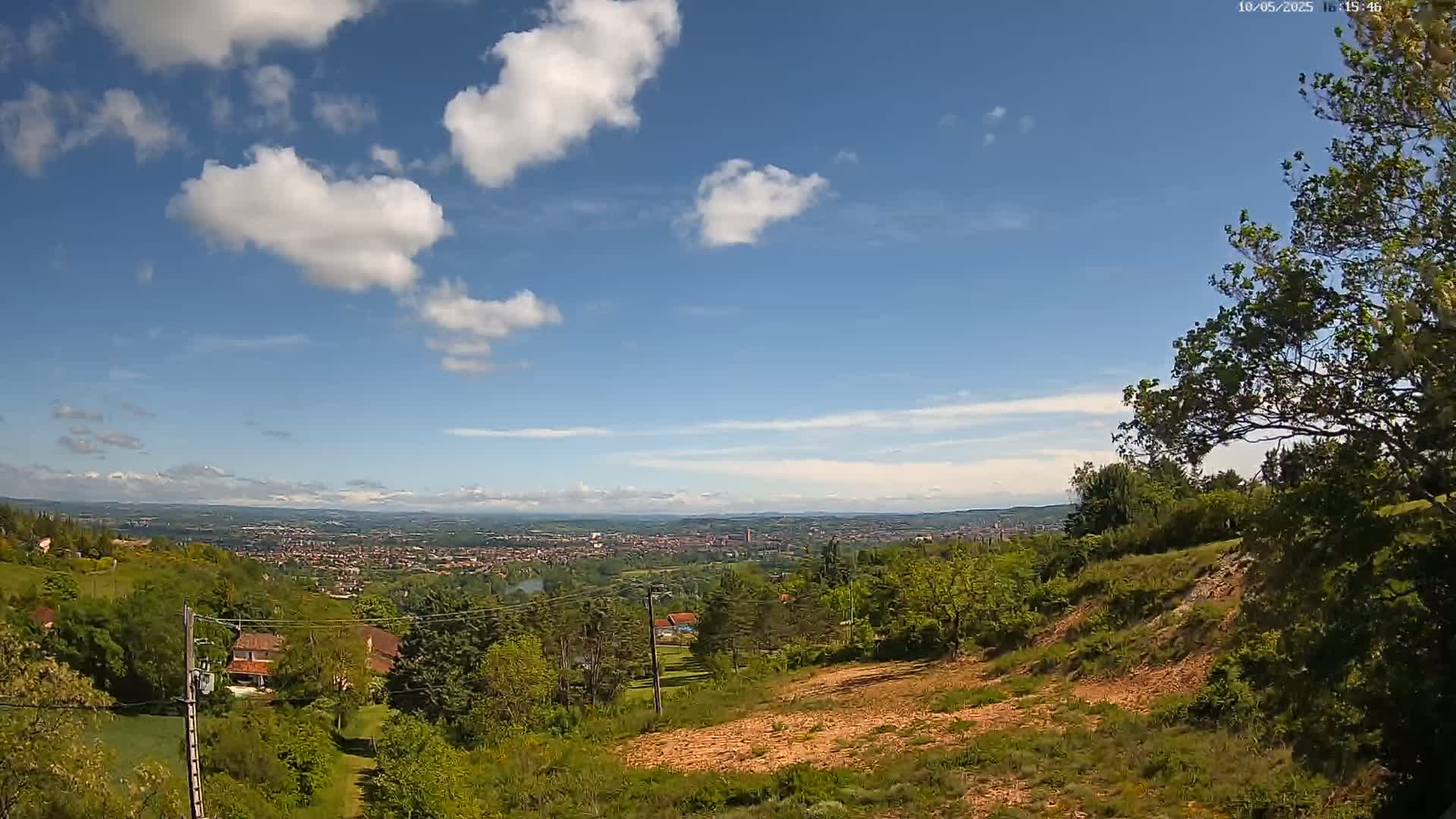 A hilly landscape with lush green vegetation overlooks a distant city under a partly cloudy blue sky.