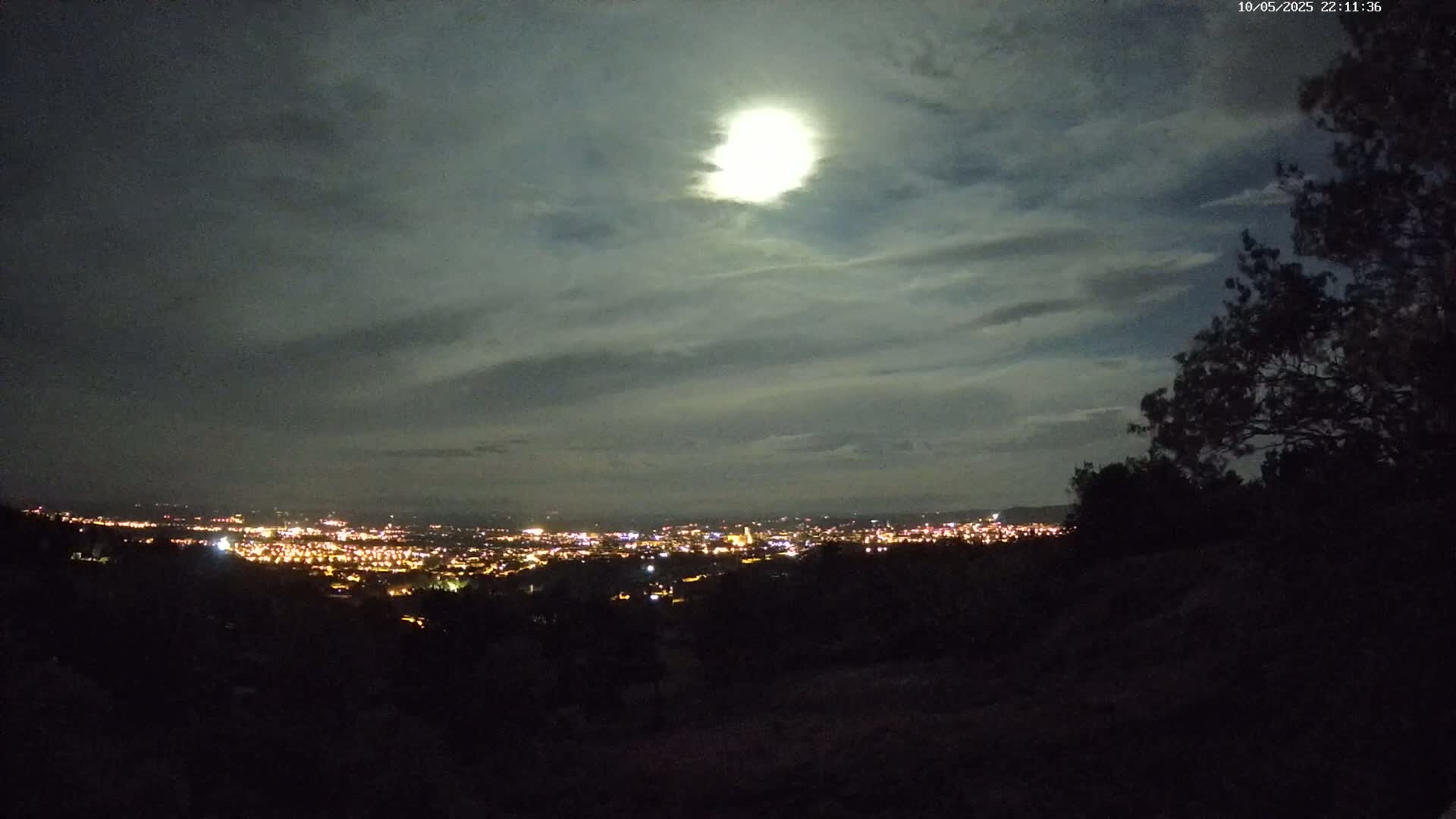 A nighttime view of a city illuminated by lights under a partly cloudy sky with a bright moon.