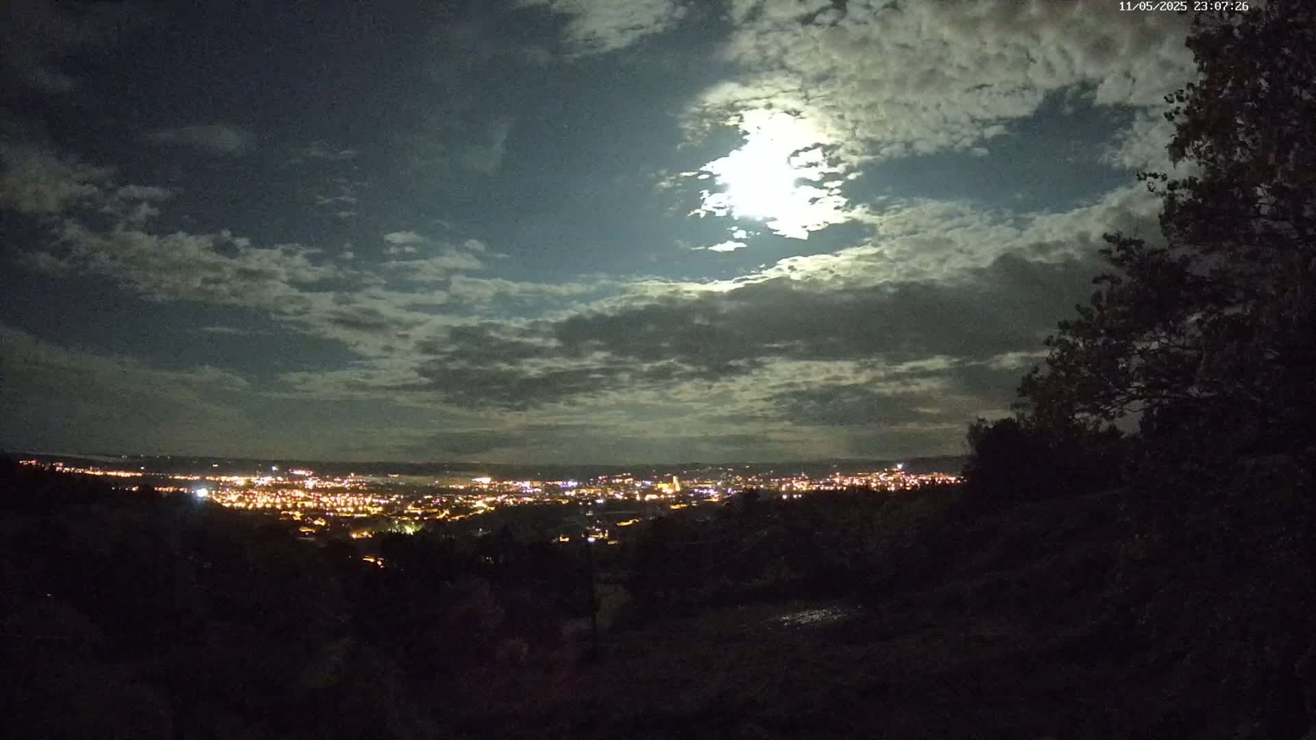 A nighttime view from a hilltop shows a city illuminated below a partly cloudy sky, with a bright moon visible through the clouds.