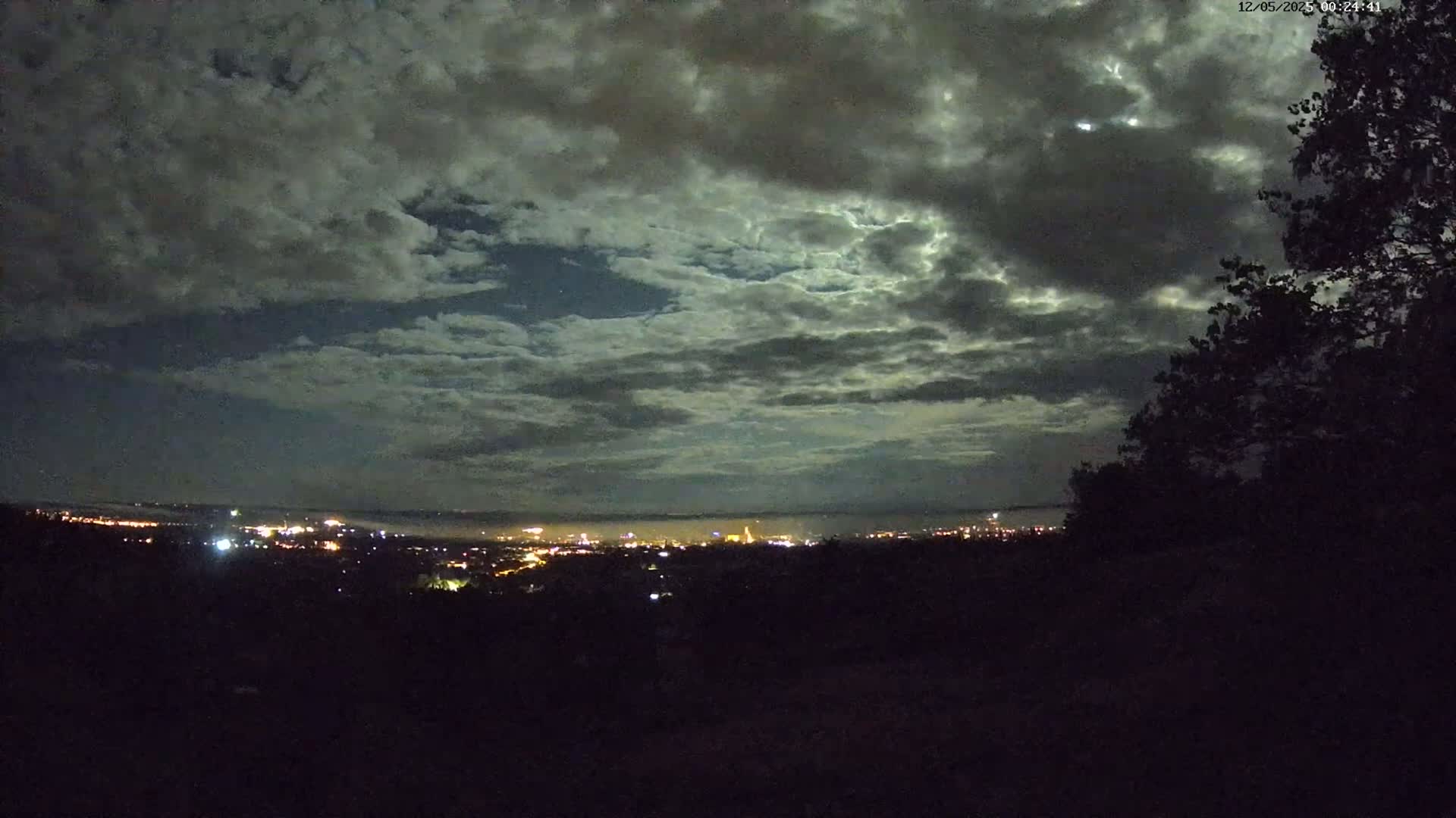 A nighttime view of a city skyline and valley illuminated by lights under a cloudy, moonlit sky.
