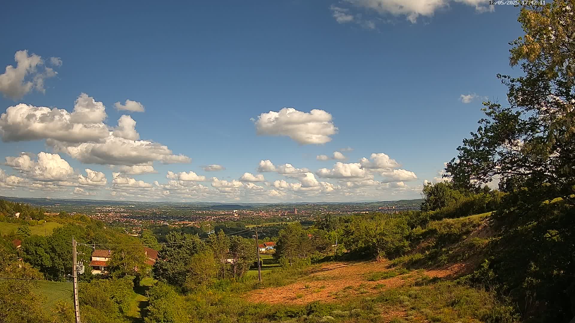 A hilly landscape with lush green vegetation and a distant view of a city under a mostly sunny sky with scattered cumulus clouds.