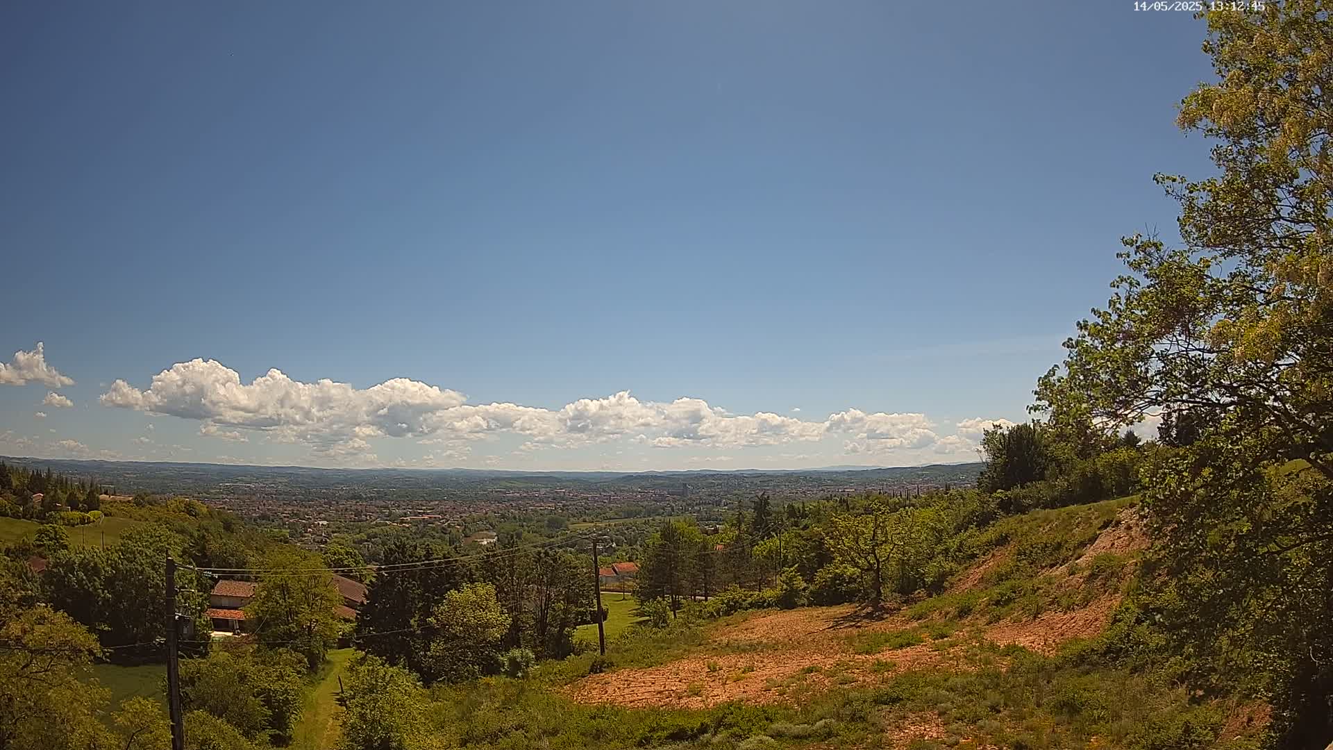 A hilly landscape with green trees and vegetation overlooks a distant city under a mostly sunny sky with a band of cumulus clouds.