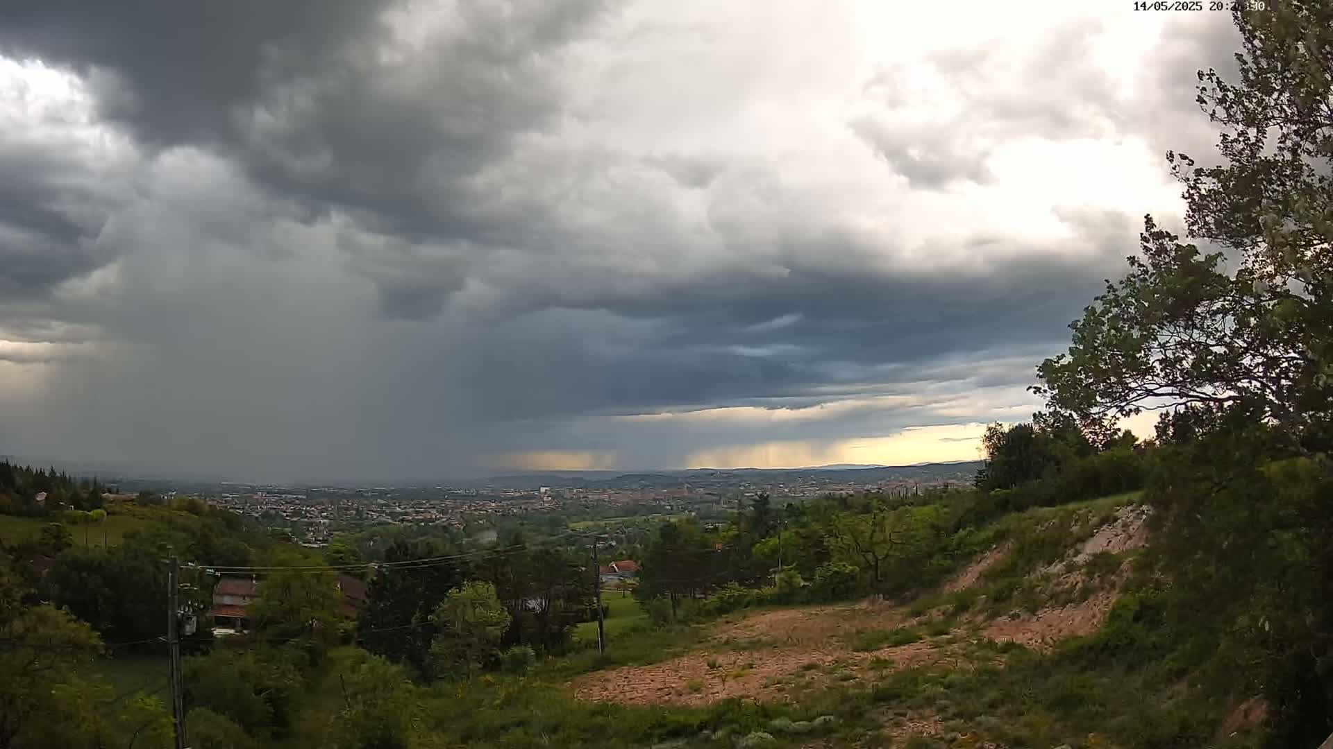 A hilly landscape with green trees and a distant city is viewed under a stormy sky with heavy rain in the distance.