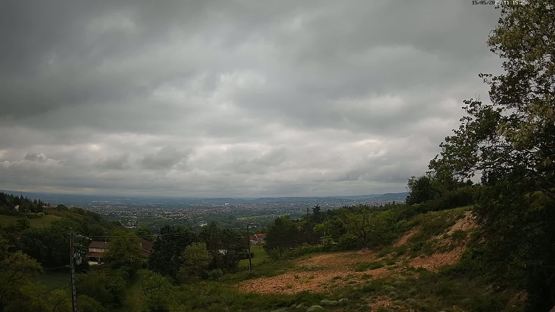 An overcast sky hangs over a hilly landscape, showing a distant cityscape nestled among green trees and a cleared, reddish-brown hillside in the foreground.