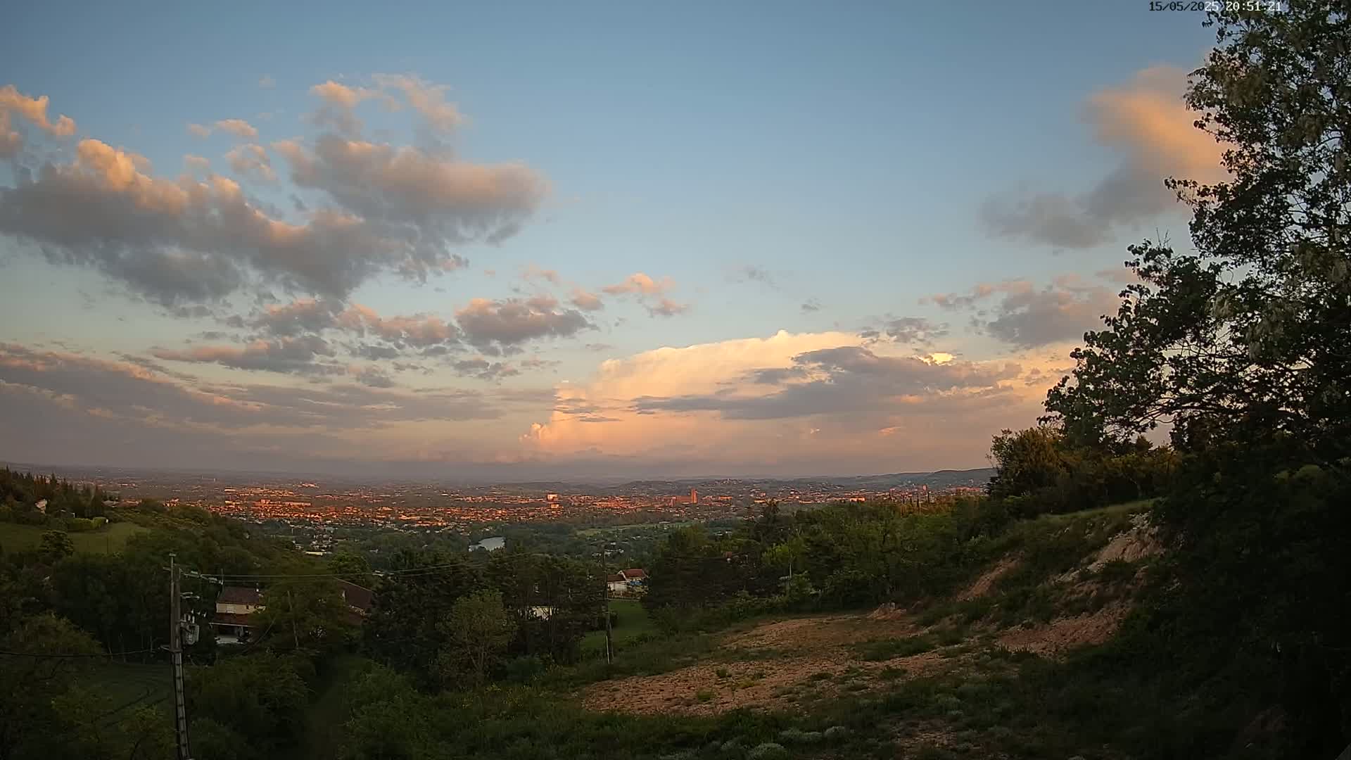 A sprawling city is visible in the distance under a partly cloudy sunset sky, viewed from a hilltop covered in lush green vegetation.