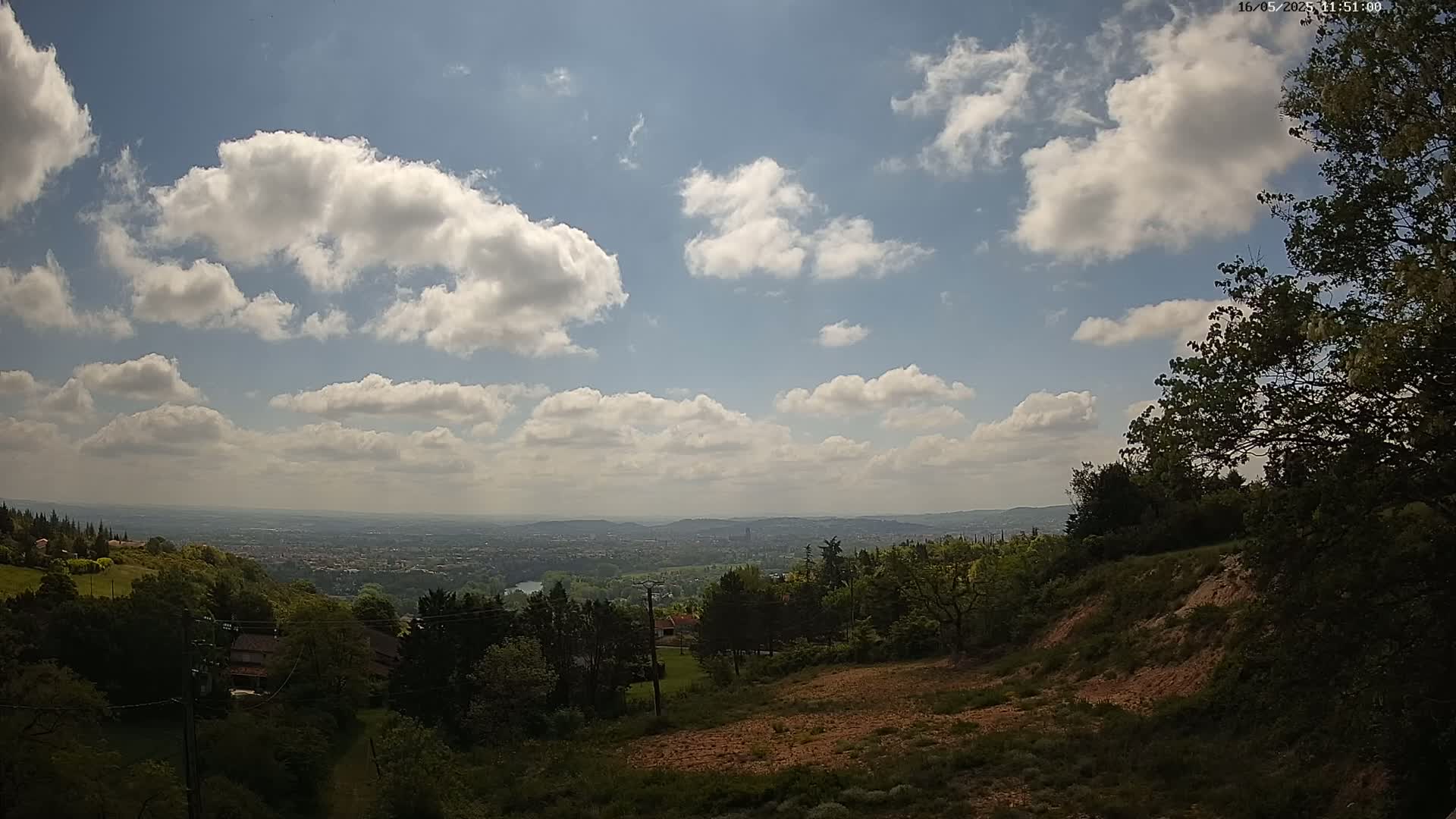 A partly cloudy sky overlooks a hilly landscape with green trees and a distant city view.