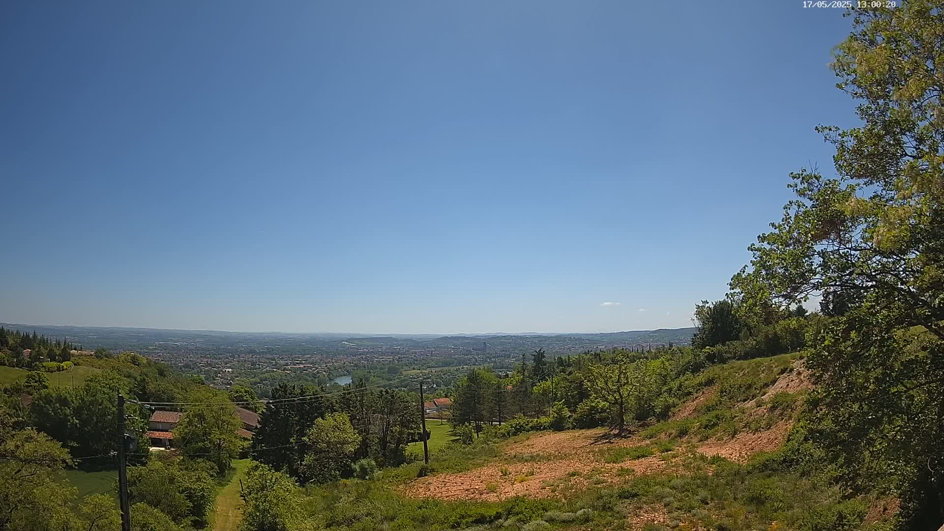 A panoramic view on a clear, sunny day shows a distant city nestled in a valley, surrounded by lush green hills and trees, with a portion of exposed, reddish-brown earth in the foreground.