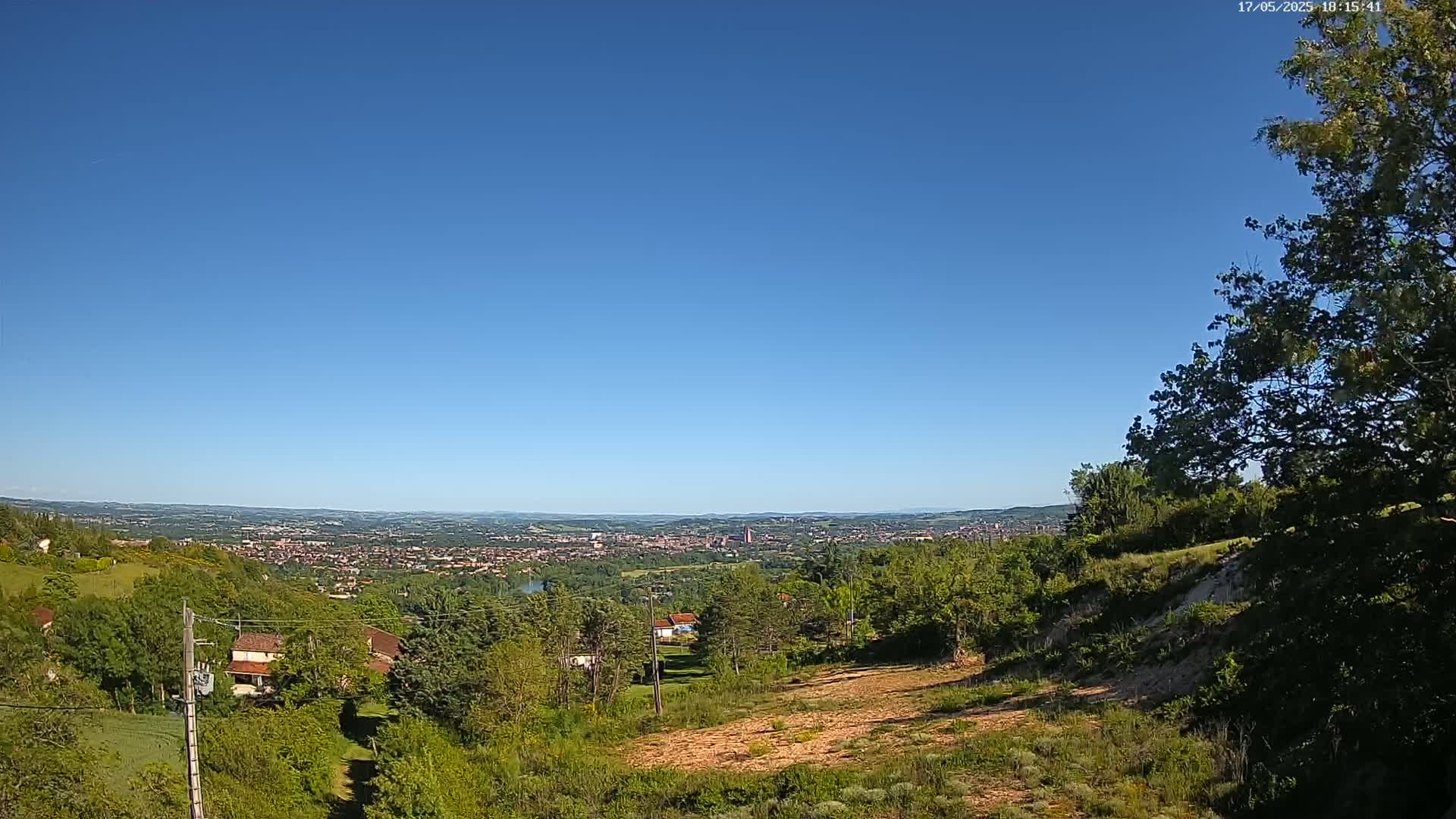 A sunny, expansive view from a hillside shows a distant city surrounded by lush green trees and vegetation.