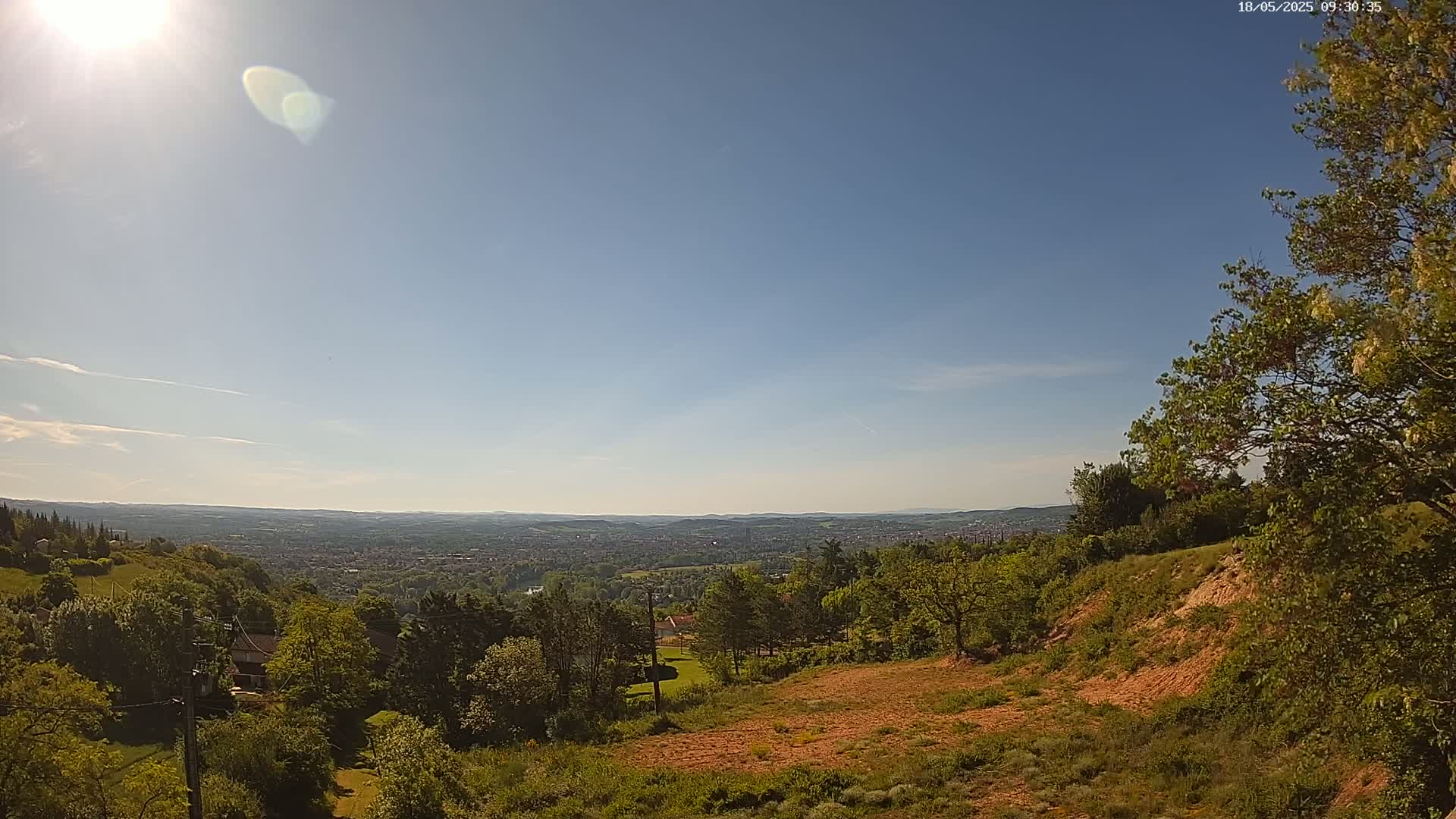 A sunny, mostly clear day reveals a panoramic view of a sprawling town nestled in a valley, surrounded by lush green hills and trees.