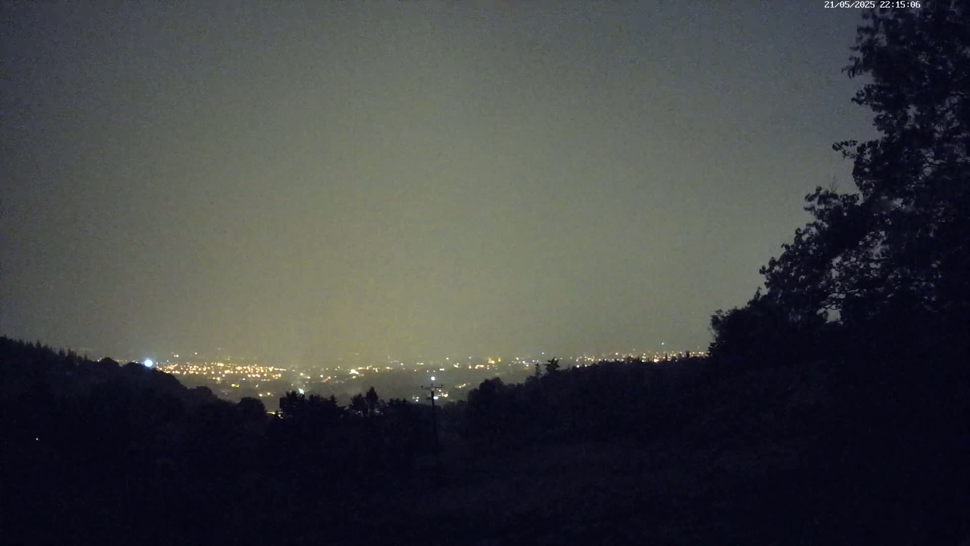 A hazy night view shows a distant city glowing with lights below a dark, silhouetted hillside.