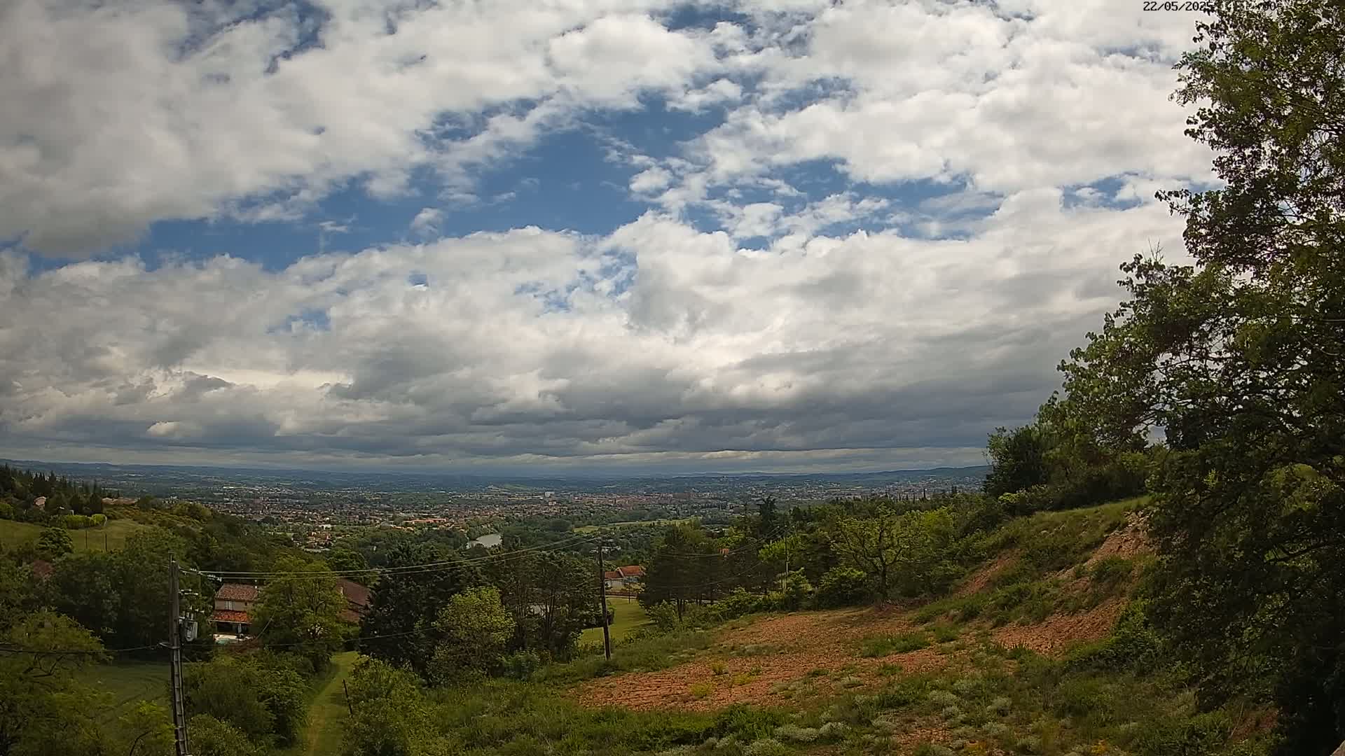 A partly cloudy sky overlooks a sprawling city visible in the distance from a hilly, wooded area.