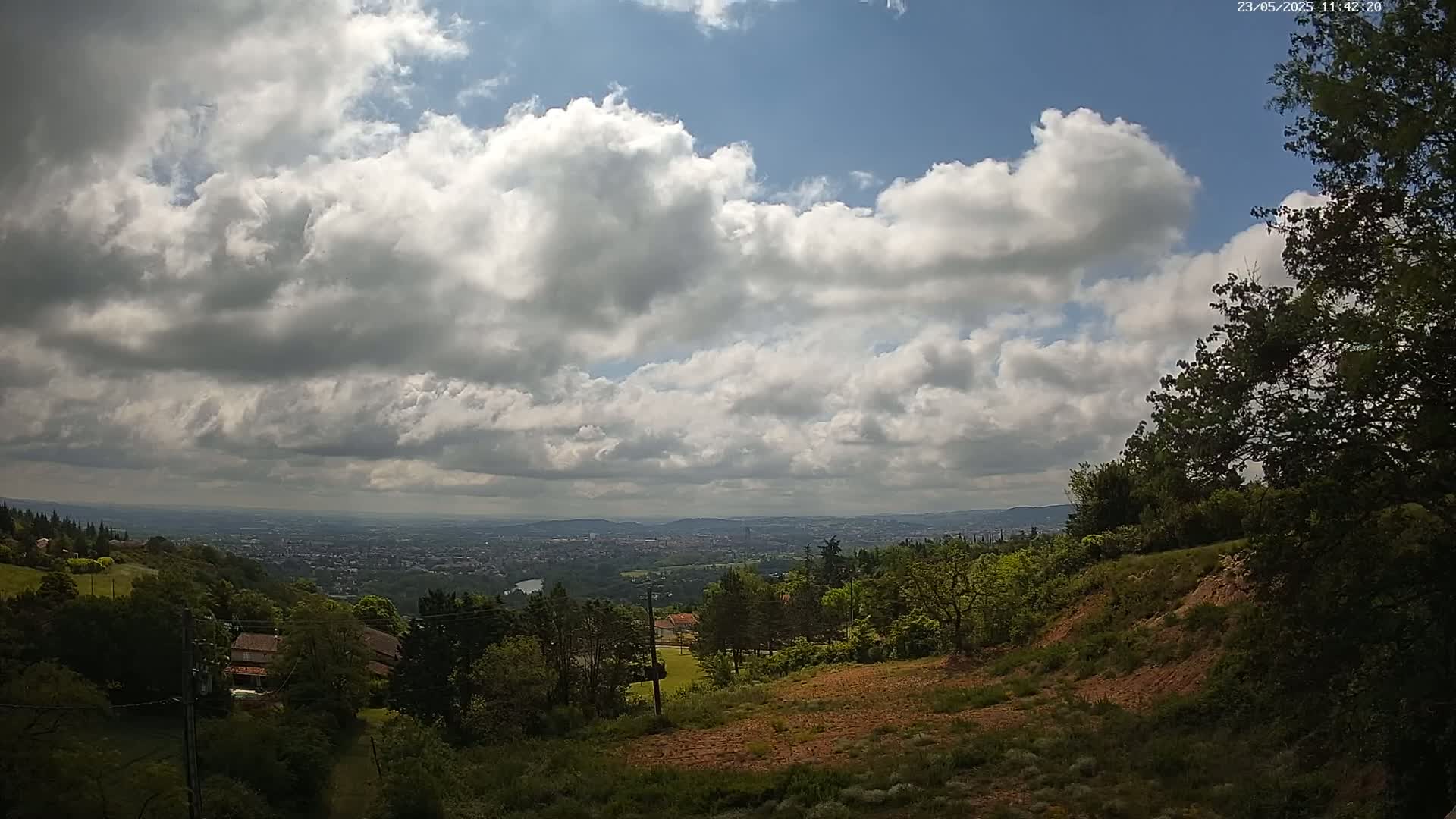 A partly cloudy sky overlooks a hilly landscape with green trees and a distant city visible in the background.