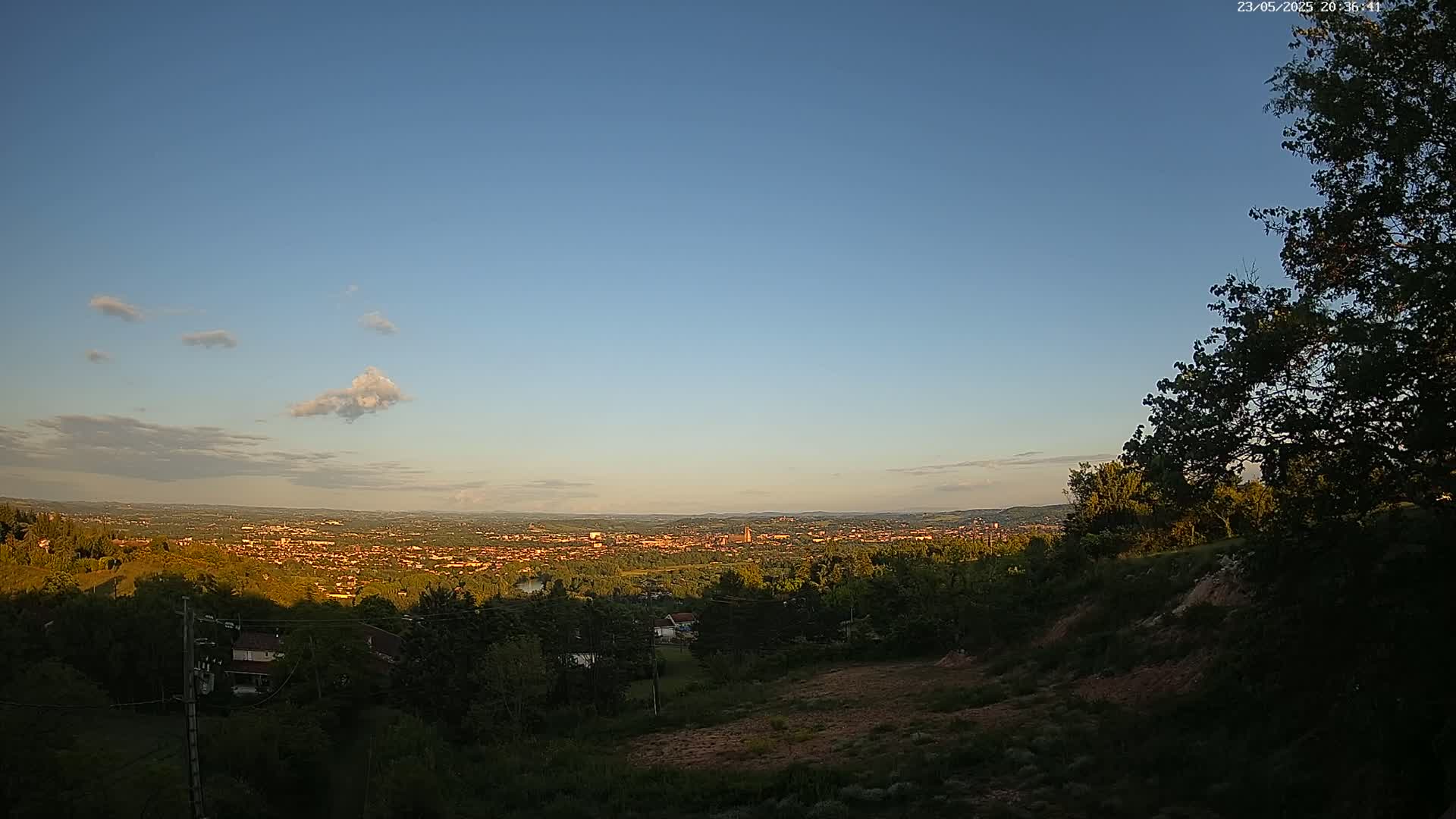 A sprawling city is visible in the distance under a mostly clear, blue sky with a few scattered clouds at sunset, viewed from a high vantage point amid trees and brush.