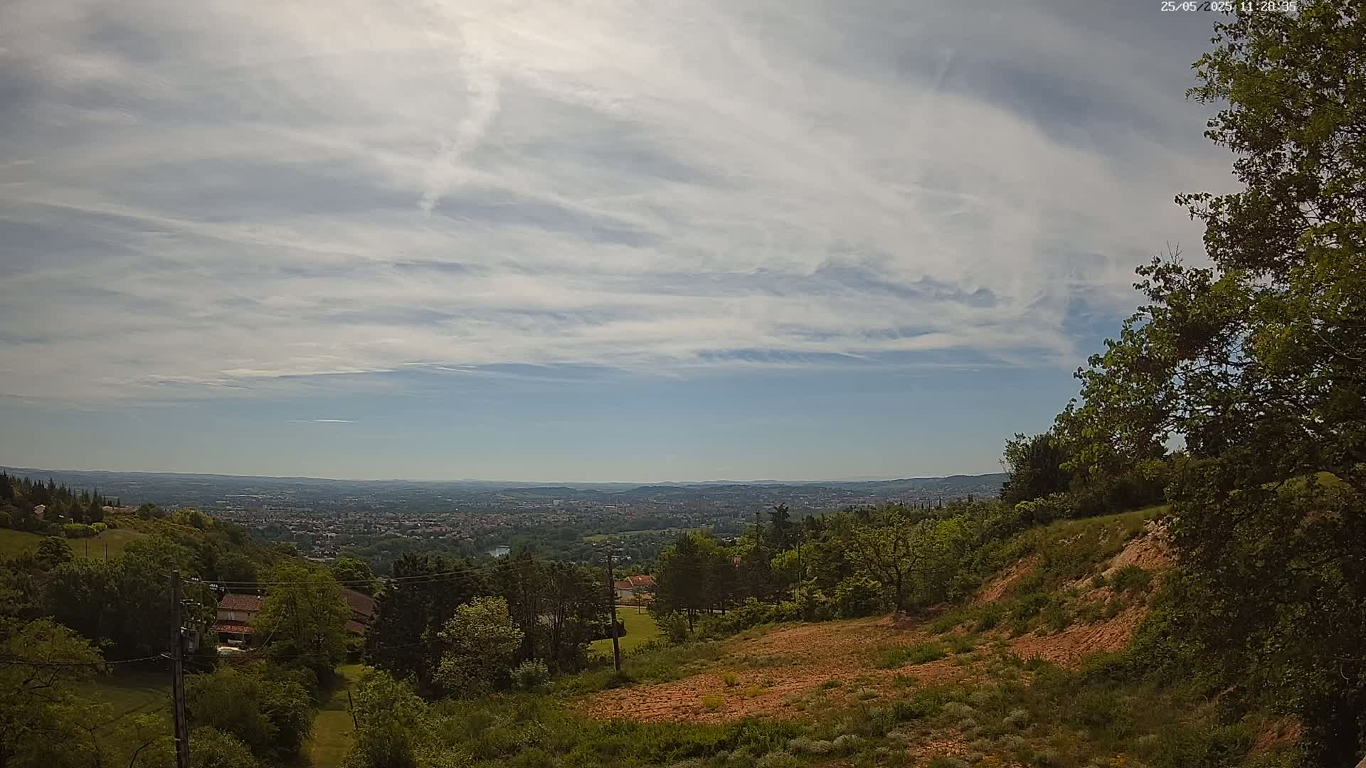 A partly cloudy sky overlooks a hilly landscape with green trees and a distant city under a bright sun.